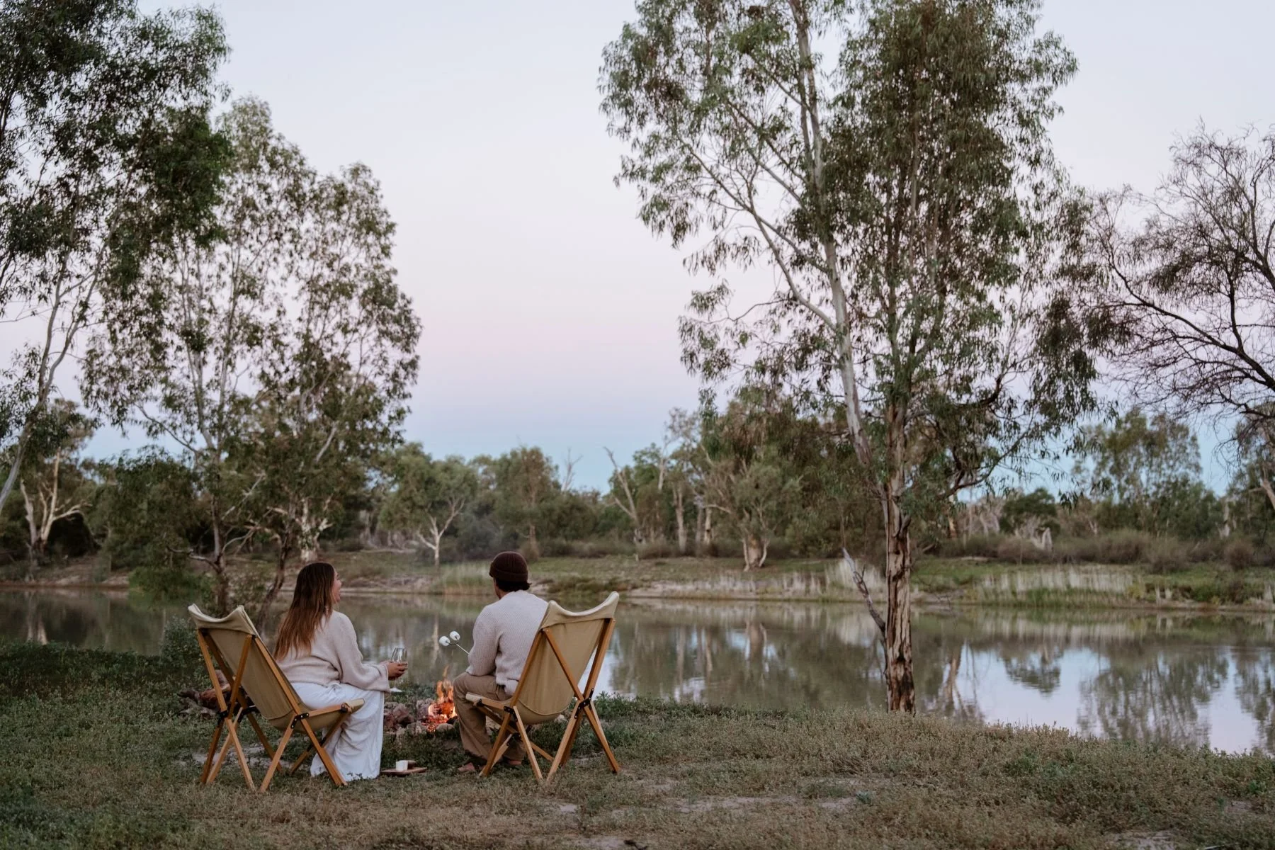 A couple sitting on chairs by a campfire with the river murray in the background, they have their backs to the camera and are facing the fire and the river scene