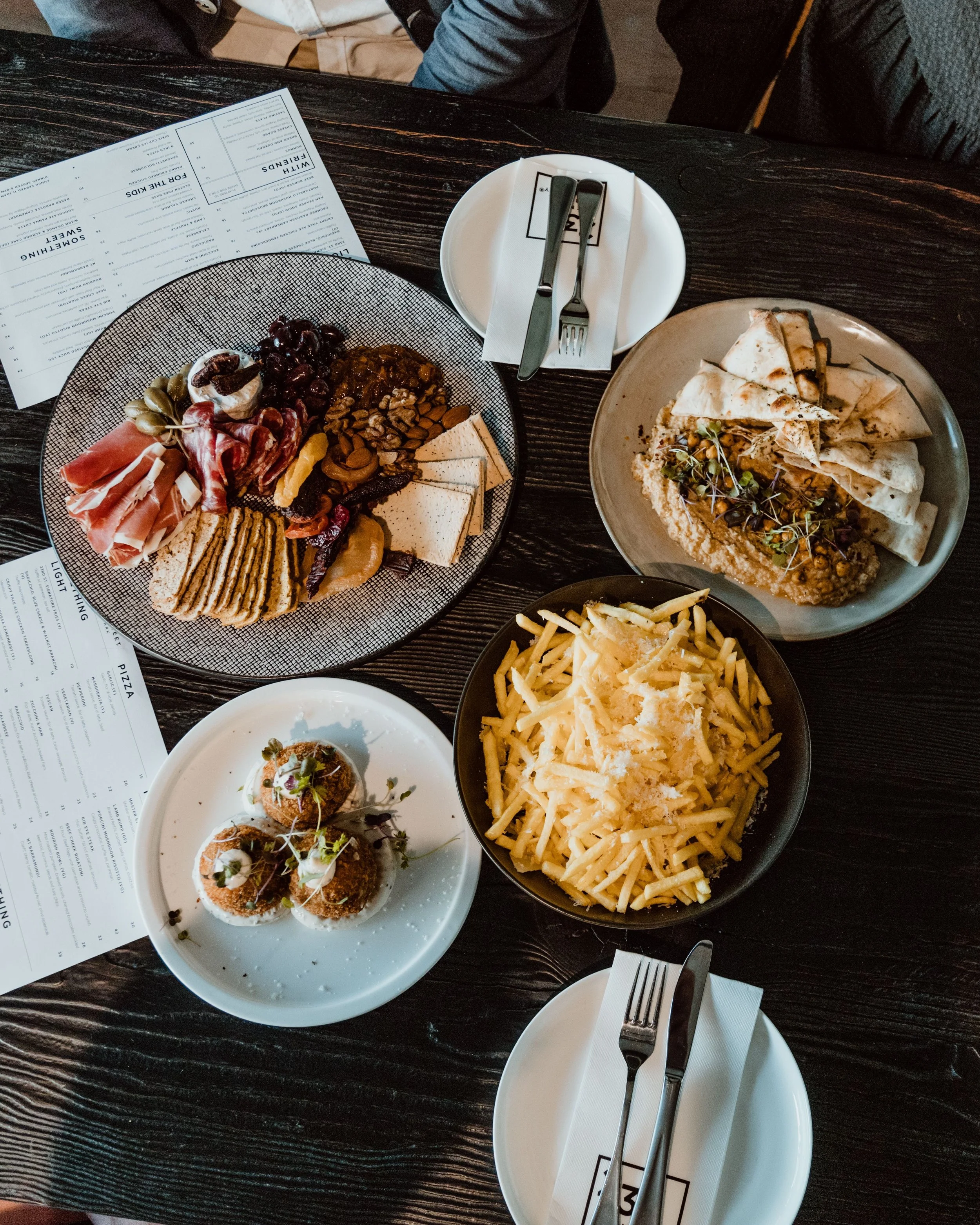 A table setting with various dishes including a charcuterie board, a plate of hummus with flatbread, a bowl of thin-cut French fries topped with cheese, and a small plate with three arancini balls. Menus and a side plate with utensils are also visible.
