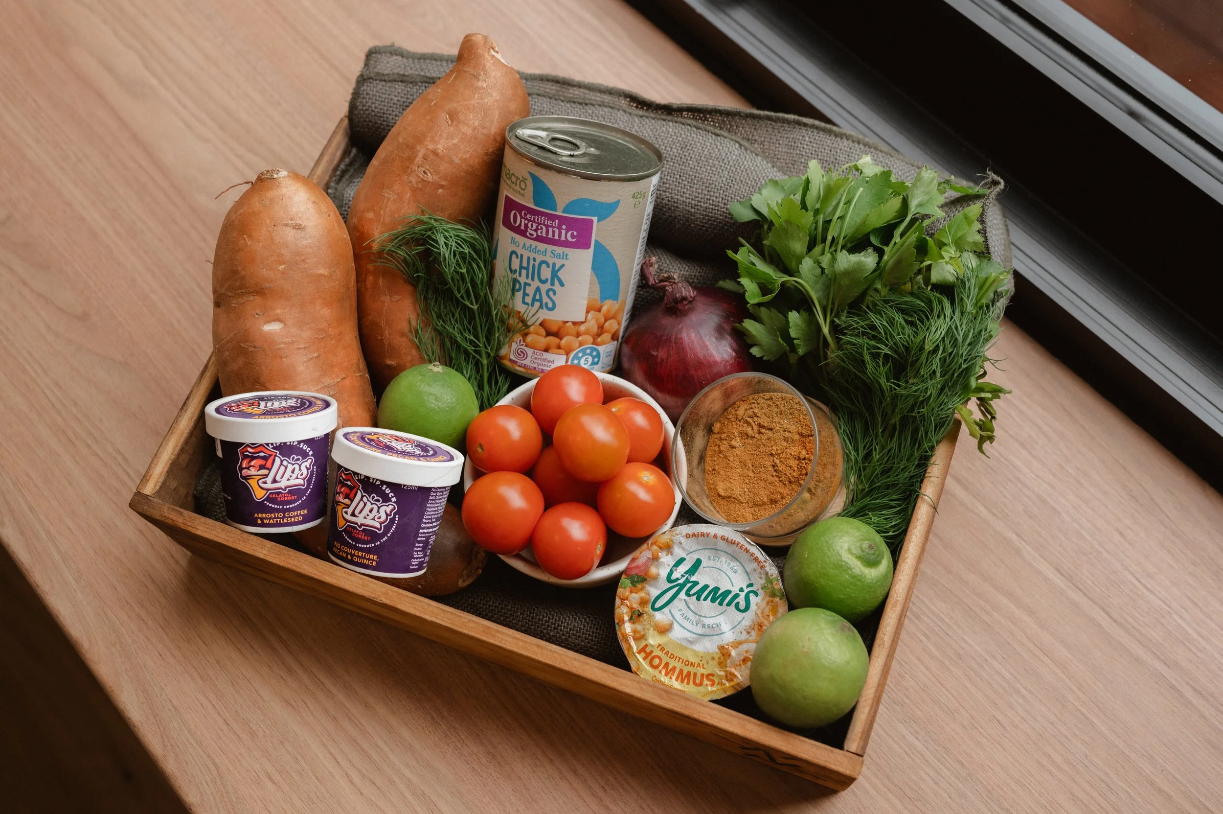 Wooden tray with sweet potatoes, organic chickpeas can, cherry tomatoes, red onion, fresh herbs, limes, yogurt cups, hummus, and a bowl of spice on a wooden surface.