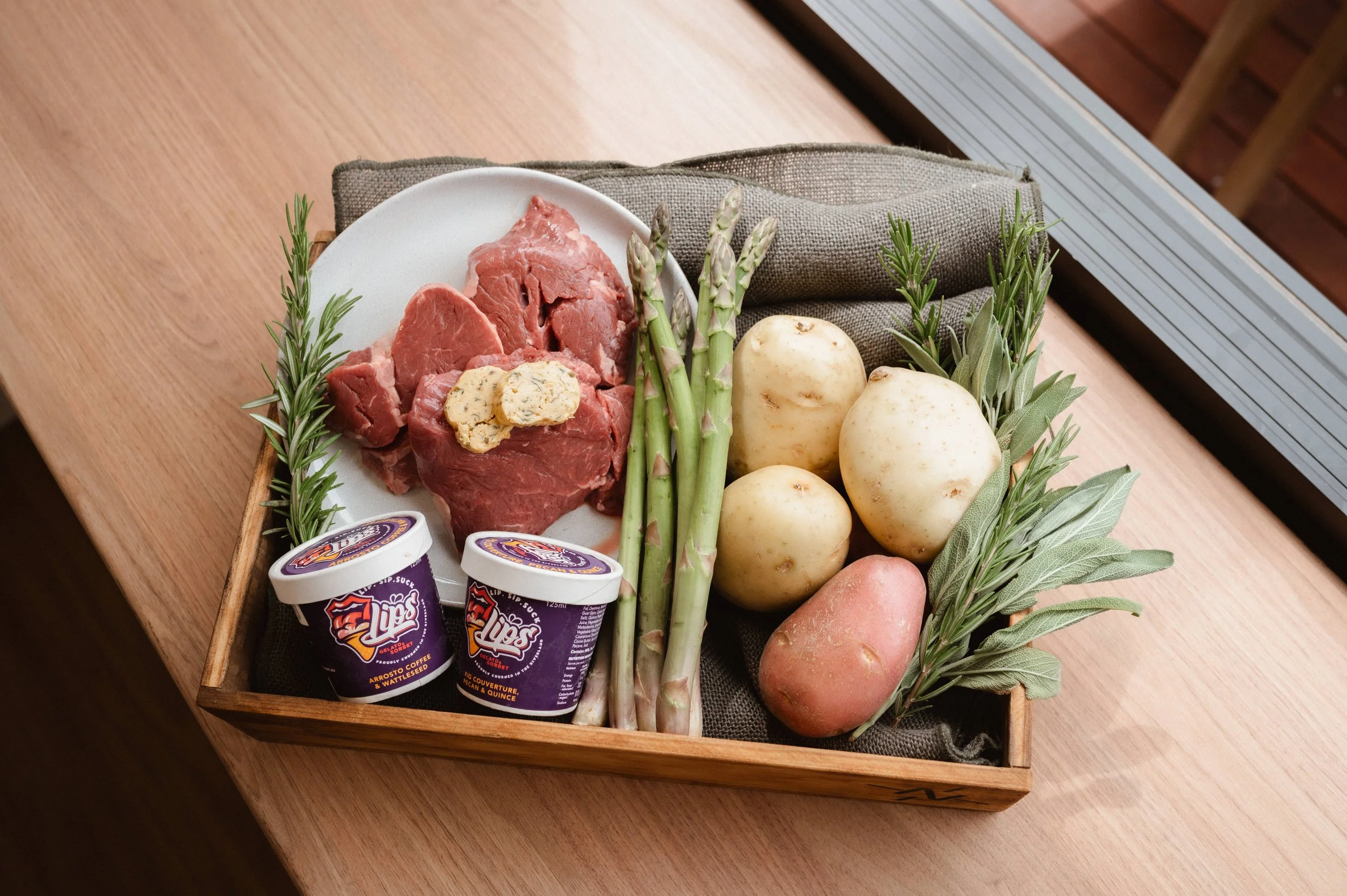 Wooden tray with raw steak, butter, asparagus, potatoes, fresh herbs, and two containers of gelato on a wooden surface.