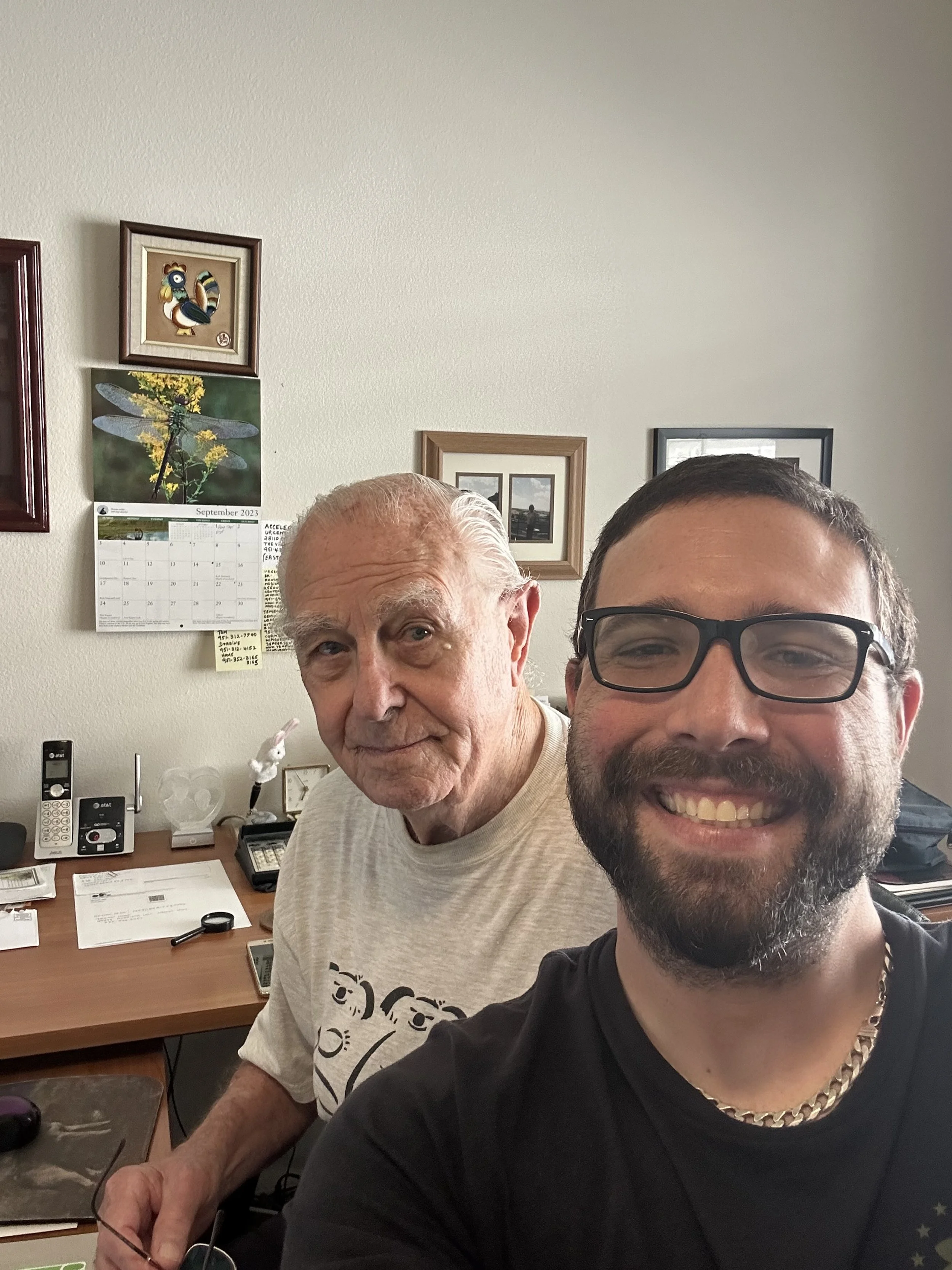 Two people smiling in an office with a desk, papers, and wall art in the background.