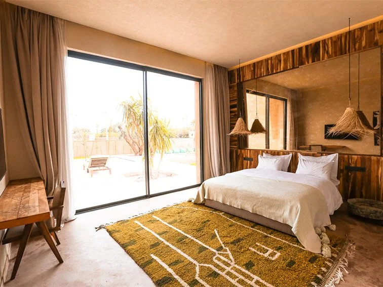 Modern bedroom with double bed, white bedding, woven lampshades, patterned rug, wooden furniture, large window with curtains, and view of a patio with a palm tree.