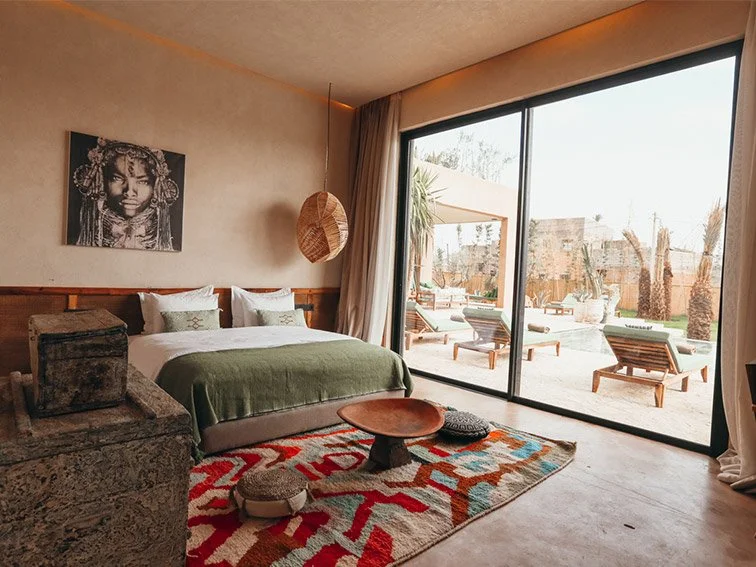 Stylish bedroom with a double bed, green blanket, decorative pillows, colorful rug, and wall art. Large glass doors offer a view of a patio with lounge chairs and palm trees.