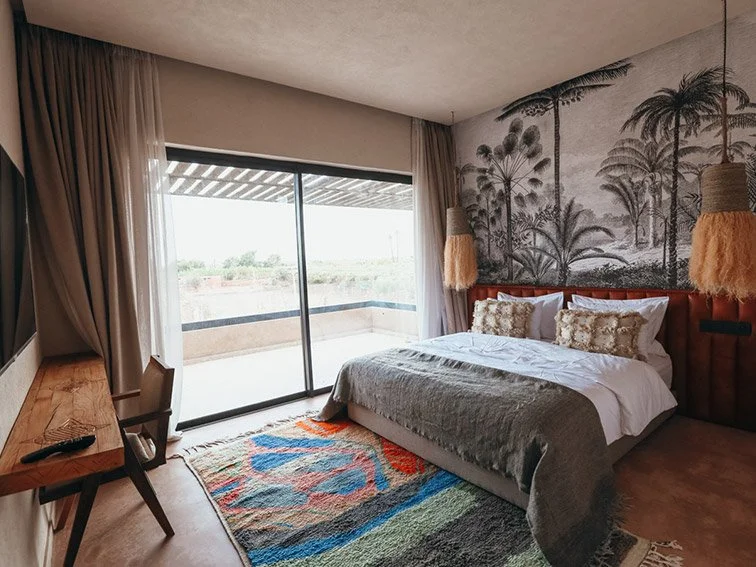 Modern bedroom with a large bed, decorative pillows, and a colorful rug. A wall mural features palm trees, and there's a wooden desk with a chair. The room has a sliding glass door leading to a patio with a view.
