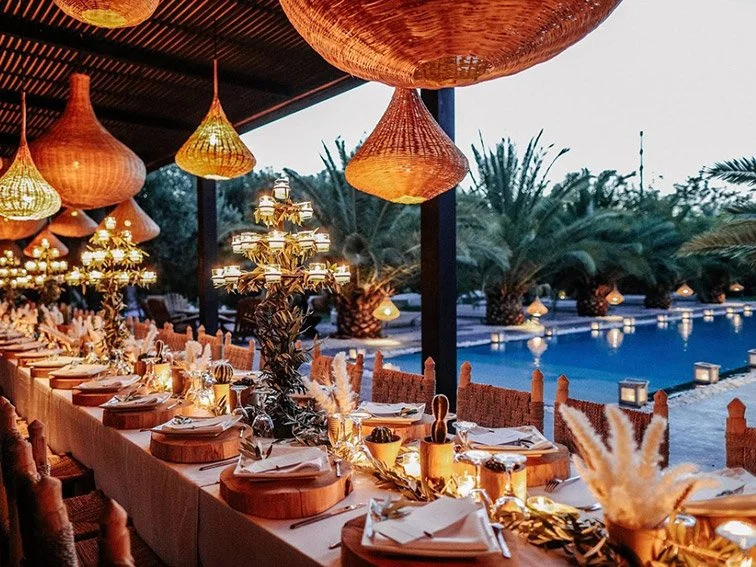 Outdoor dining setup with elegant table settings, candlelit decorations, and wicker lamps, near a pool with palm trees in the background.