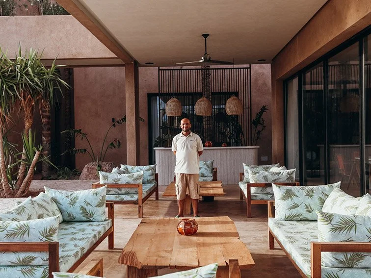 Man standing in a tropical-themed outdoor patio with wooden furniture and palm leaf cushions, beside a bar with hanging lamps.