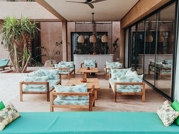 Outdoor patio seating area with wooden furniture and tropical cushions, featuring a bar with hanging lamps, plants, and decorative elements.