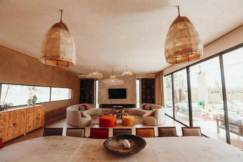 Cozy living room with curved sofa, colorful cushions, and large pendant lights. Features include a flat-screen TV, wooden sideboard, large windows, and glass doors opening to a patio.