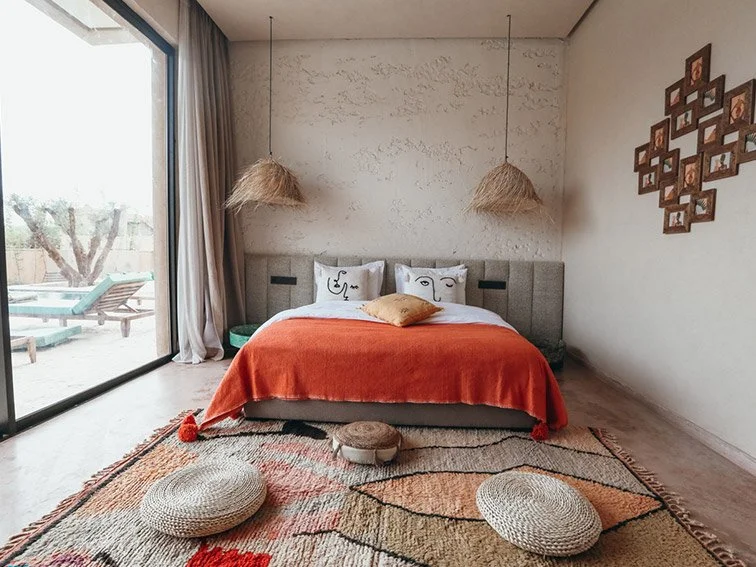 Modern bedroom with a double bed, orange bedspread, abstract pillows, and woven lamps. Large window with a view of a patio and pool. Geometric rug and decorative wall frames.