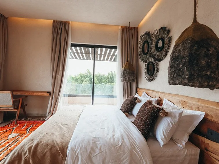 Cozy bedroom with a large bed, wooden headboard, and decorative pillows. The room features a sliding glass door with curtains, leading to a view of greenery. Two large woven pendant lights hang from the ceiling, and artistic wall decor is present. A