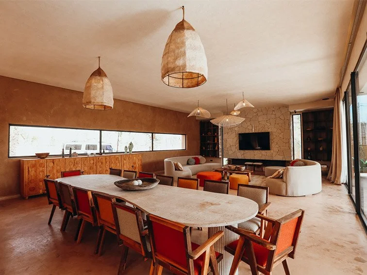 Modern living and dining room with large table, colorful chairs, pendant lighting, and a seating area with a TV on a stone wall.