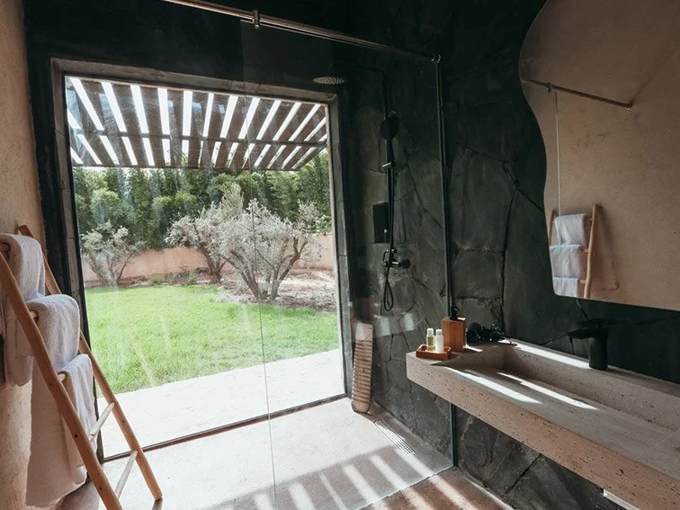Modern outdoor bathroom with a large glass wall overlooking a garden with trees and bushes, stone walls, wooden towel rack, stone sink, and natural light streaming in.