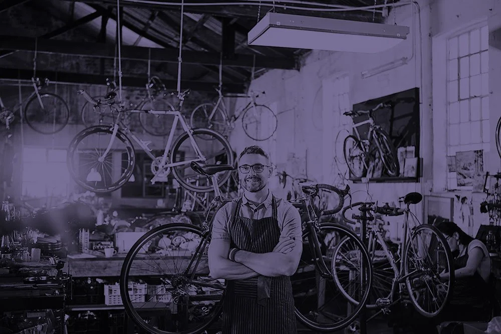 A male bike repair shop owner with glasses and wearing overalls standing in front of a collection of bikes in a bike repair shop