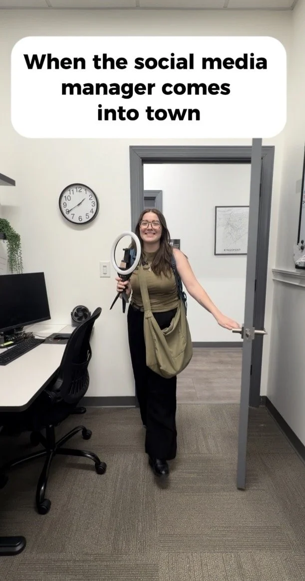 A woman smiling while holding a ring light and a phone in an office, standing in an open door with a caption above that reads 'When the social media manager comes into town'.