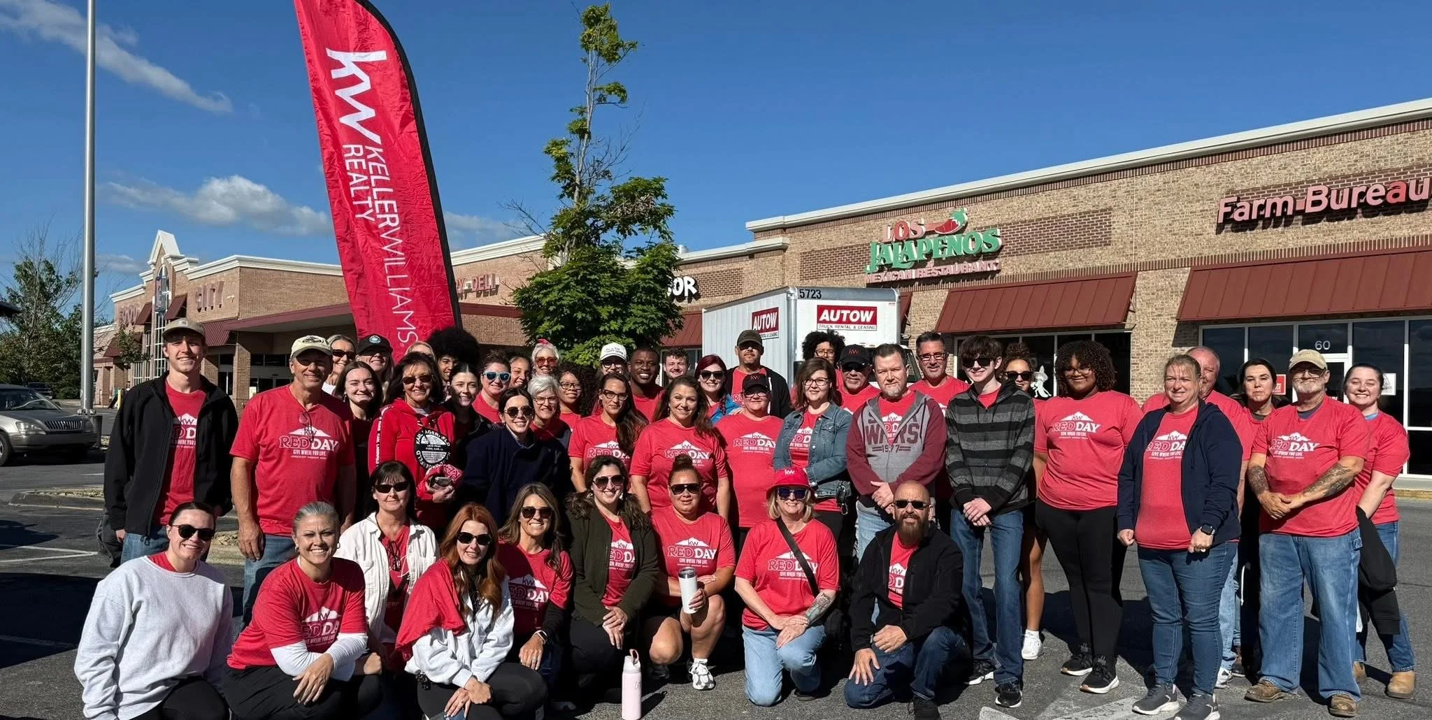 A large group of Keller Williams agents in red shirts with a 'Red Day' logo, gathered outside a shopping center.