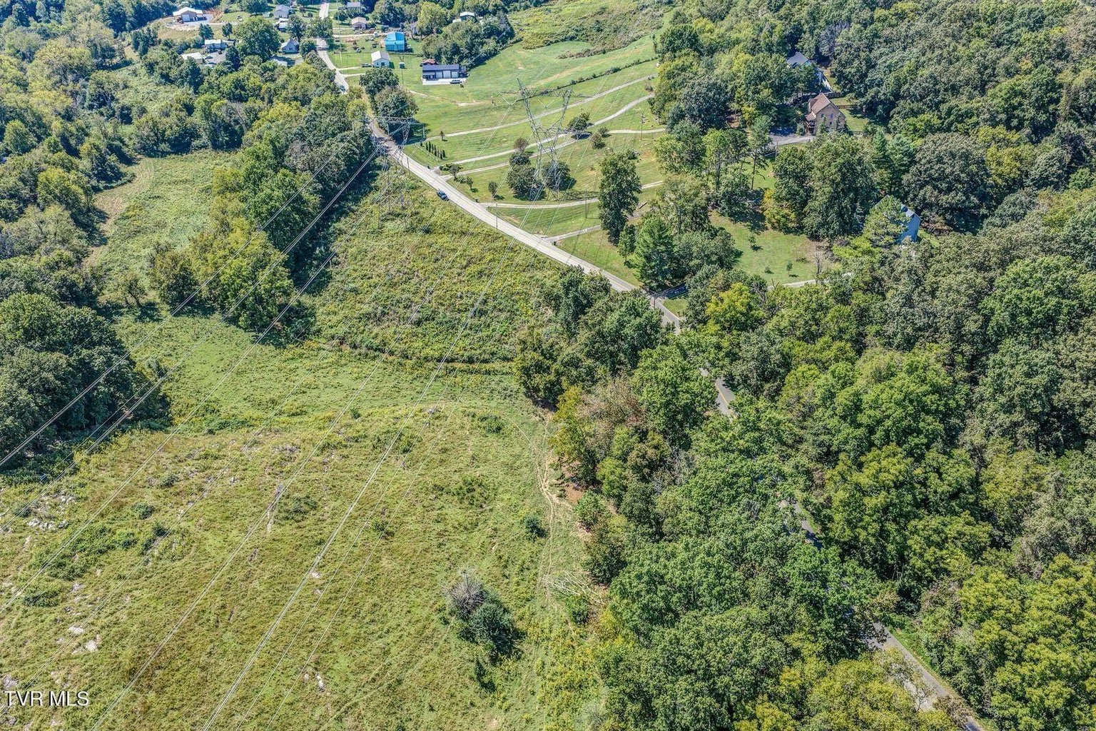 A scenic aerial view of a wooded hillside with scattered houses, a winding road, and power lines.