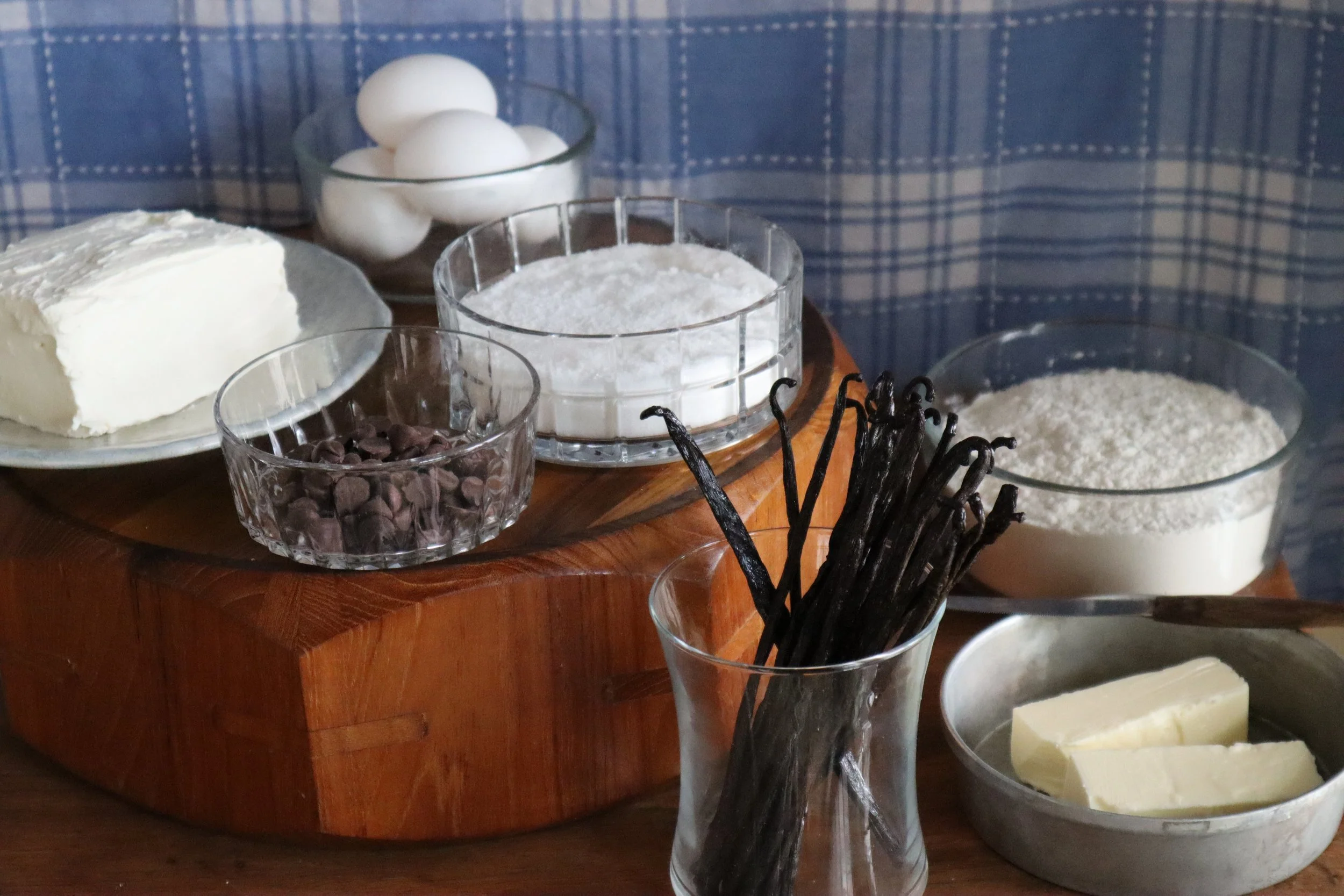 Various baking ingredients on a wooden surface, including a loaf of butter, eggs, flour, chocolate chips, vanilla pods, and a block of butter.