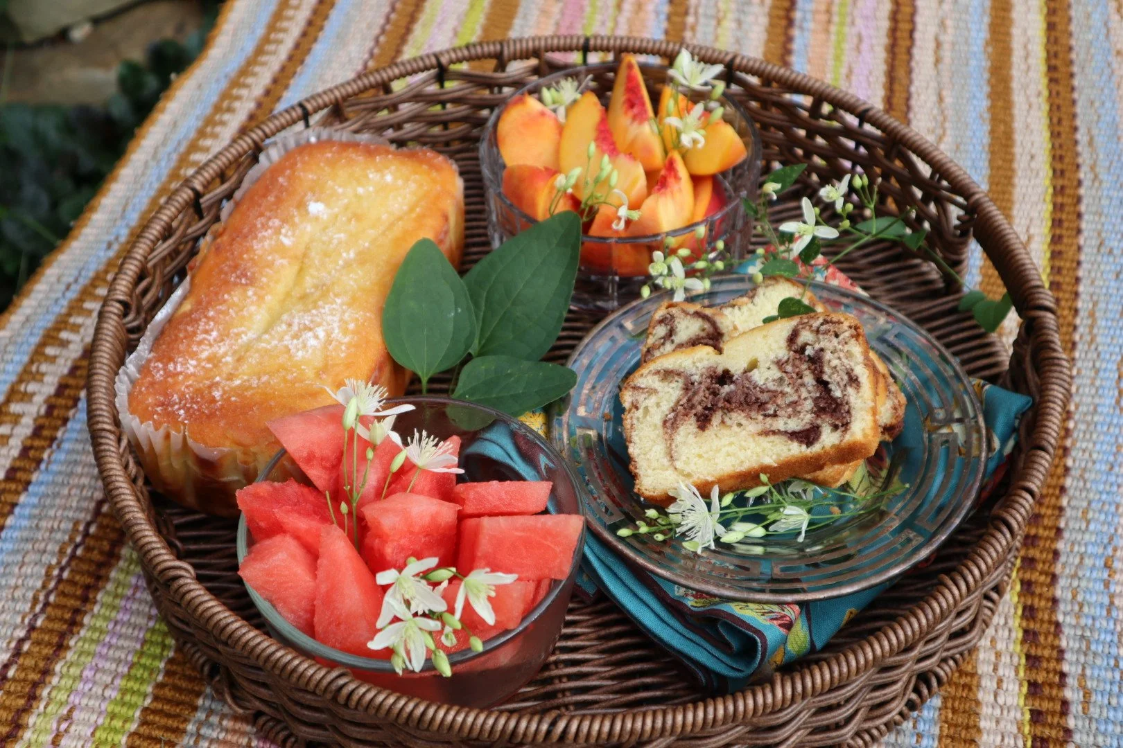 A wicker basket with slices of marble cake, chopped watermelon, a loaf cake, and sliced peaches with small white flowers. The basket is on a colorful striped tablecloth.