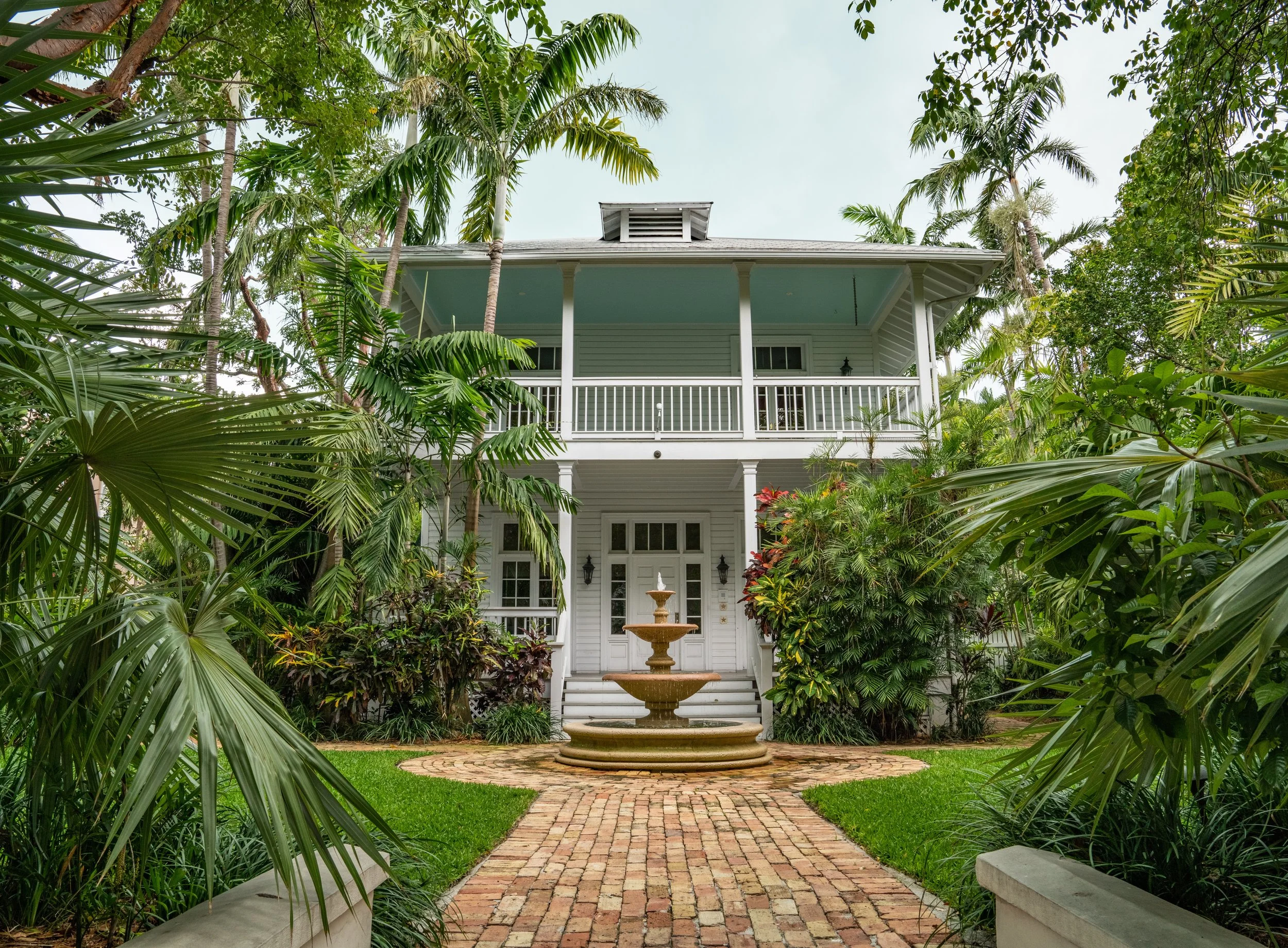 White two-story house with a front porch, surrounded by tropical plants and trees, featuring a central fountain on a brick pathway.