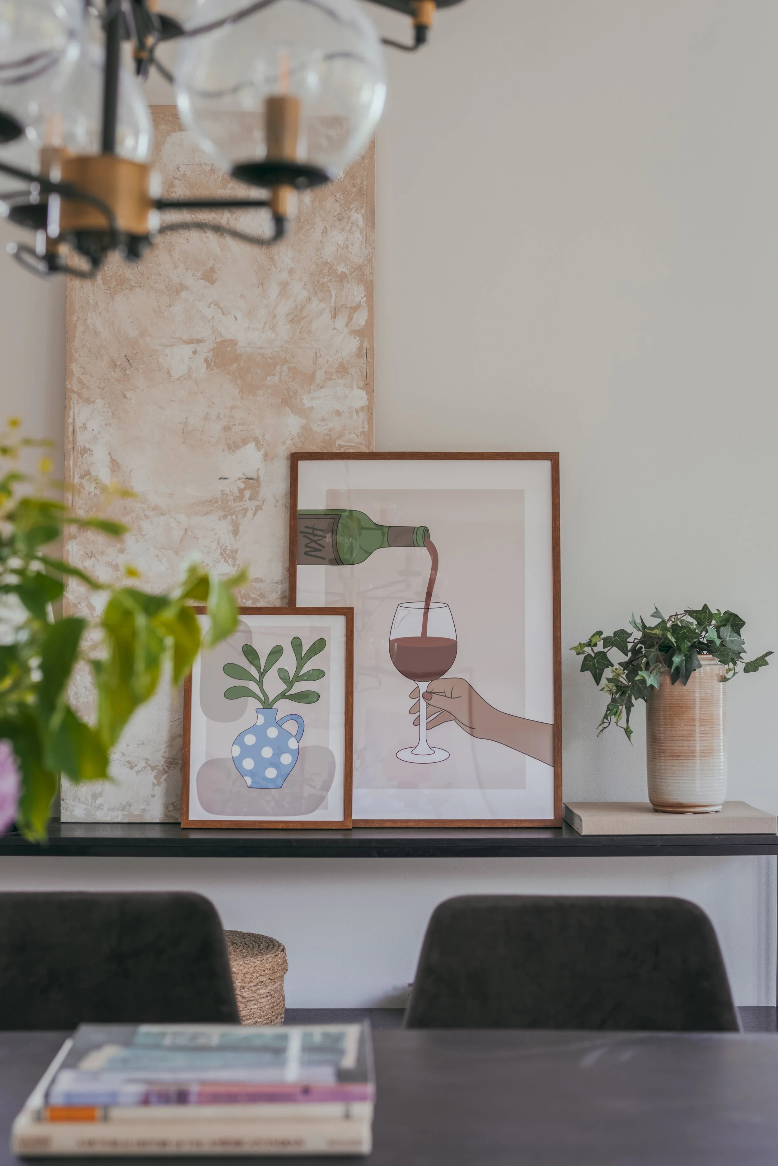 Interiörbild av ett vardagsrum eller ett café med konstverk av illustrationsbilder av växter i vaser och ett glas vin med en hand som håller ett vinprov, växter i en vas, vägg med konstverket och ett grönt växtblad i förgrunden.