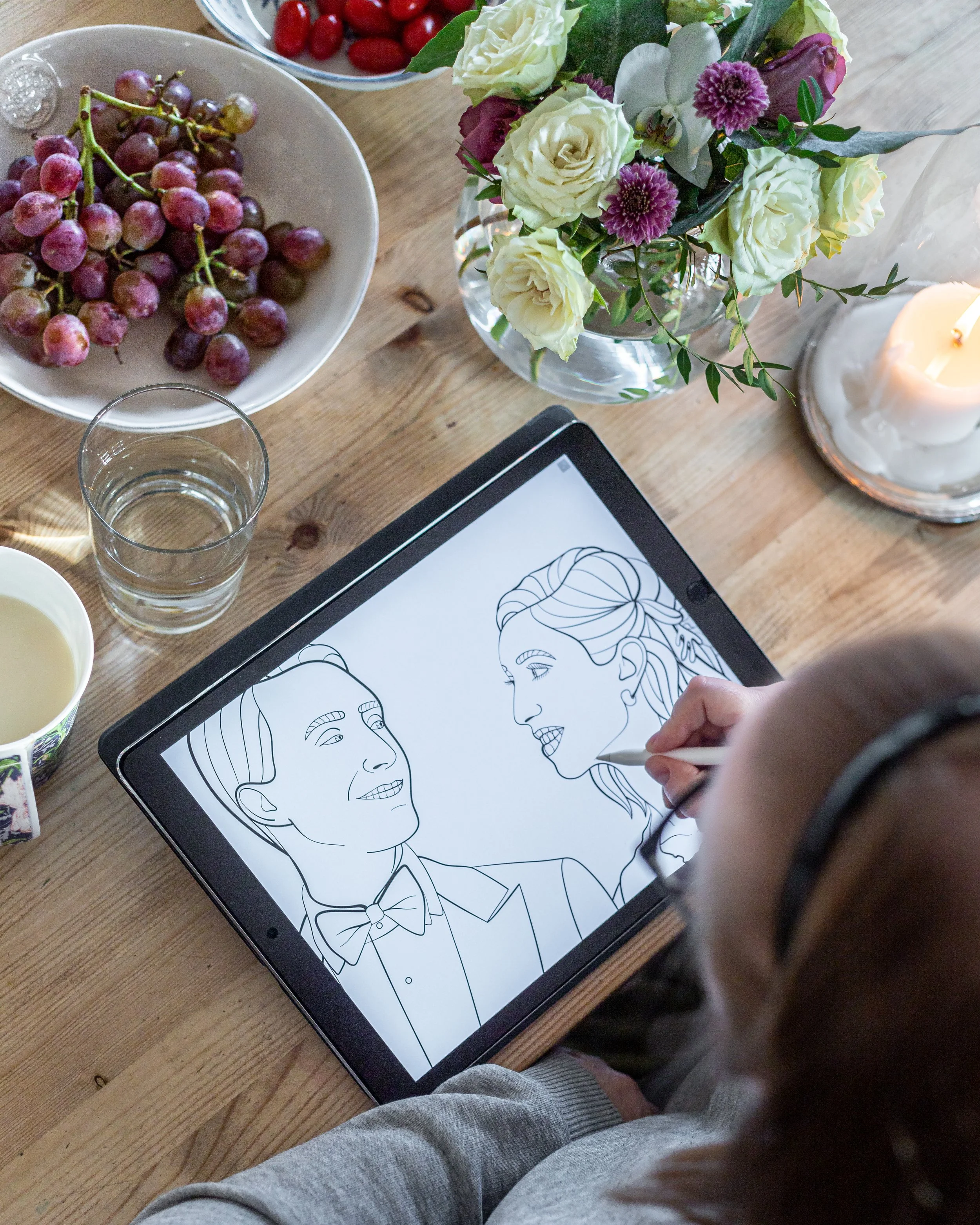 Person ritar porträtt av två kvinnor på en surfplatta, omgivande av ett bord med frukt, blommor och ett ljus.