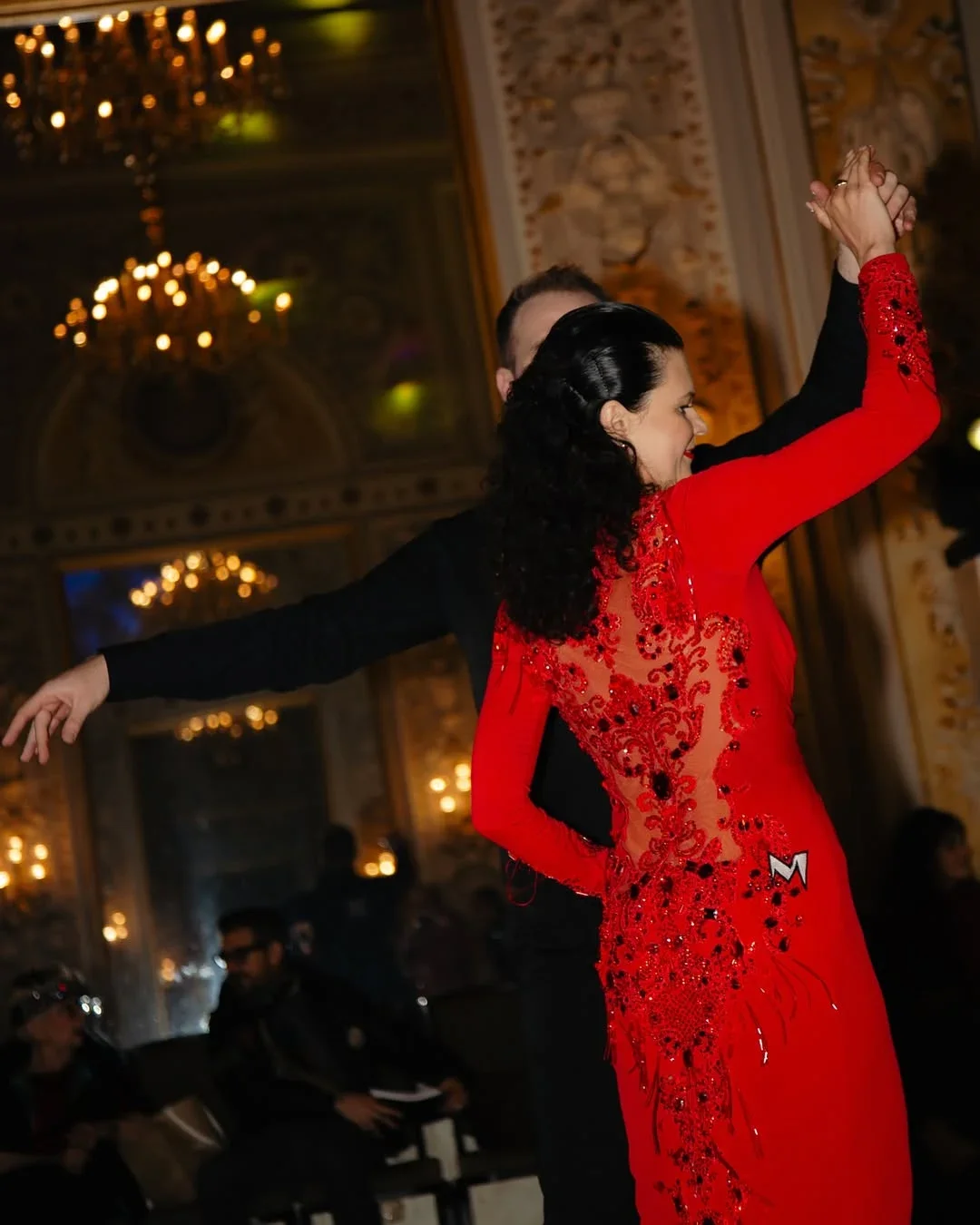 Couple performing a ballroom dance at a competition in an elegant ballroom with crystal chandeliers, the woman wearing a red beaded gown