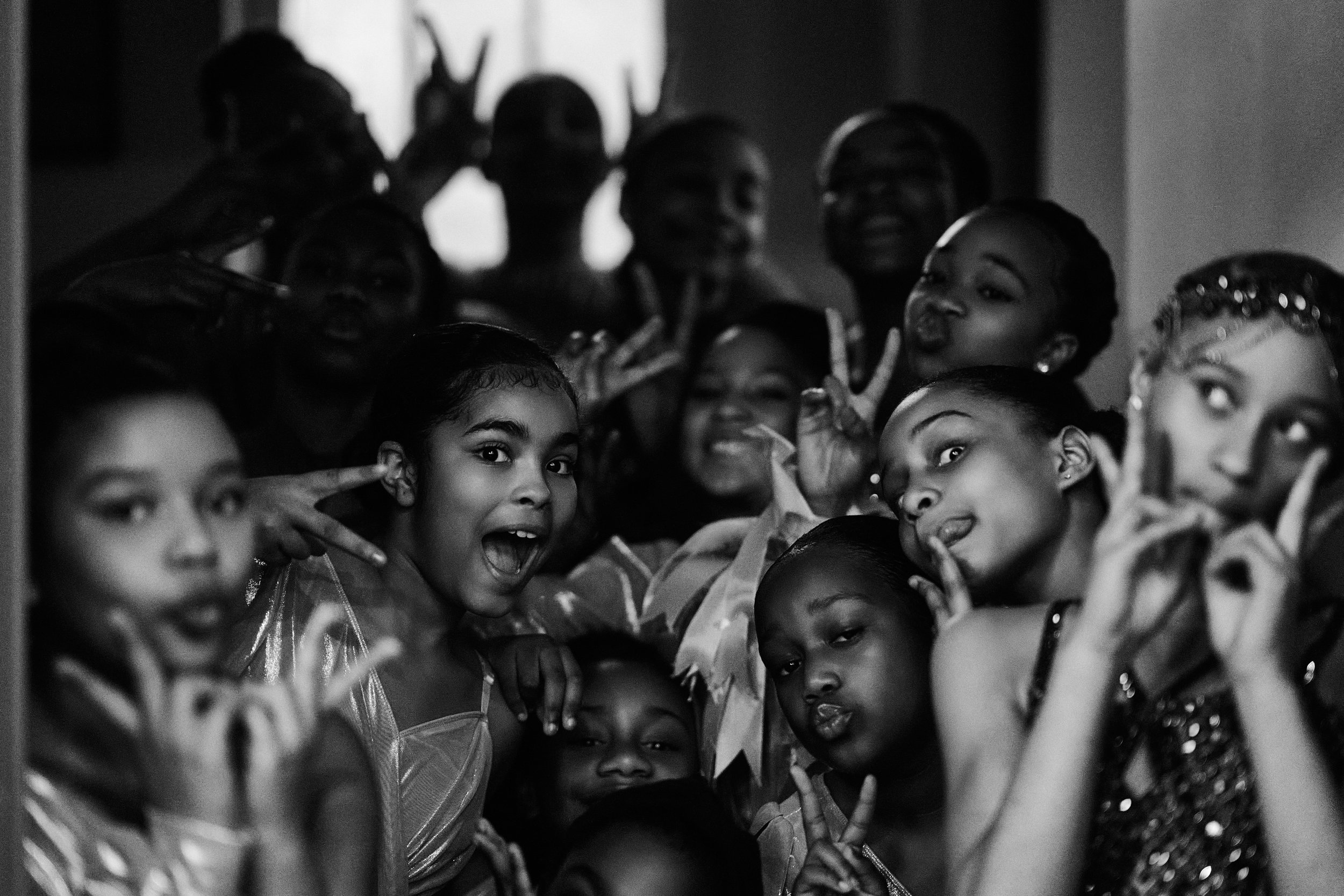 A group of young girls with some smiling and making peace signs, dressed in costumes, possibly for a dance or performance, with some in the front and others in the back, inside a room with light coming through the window.