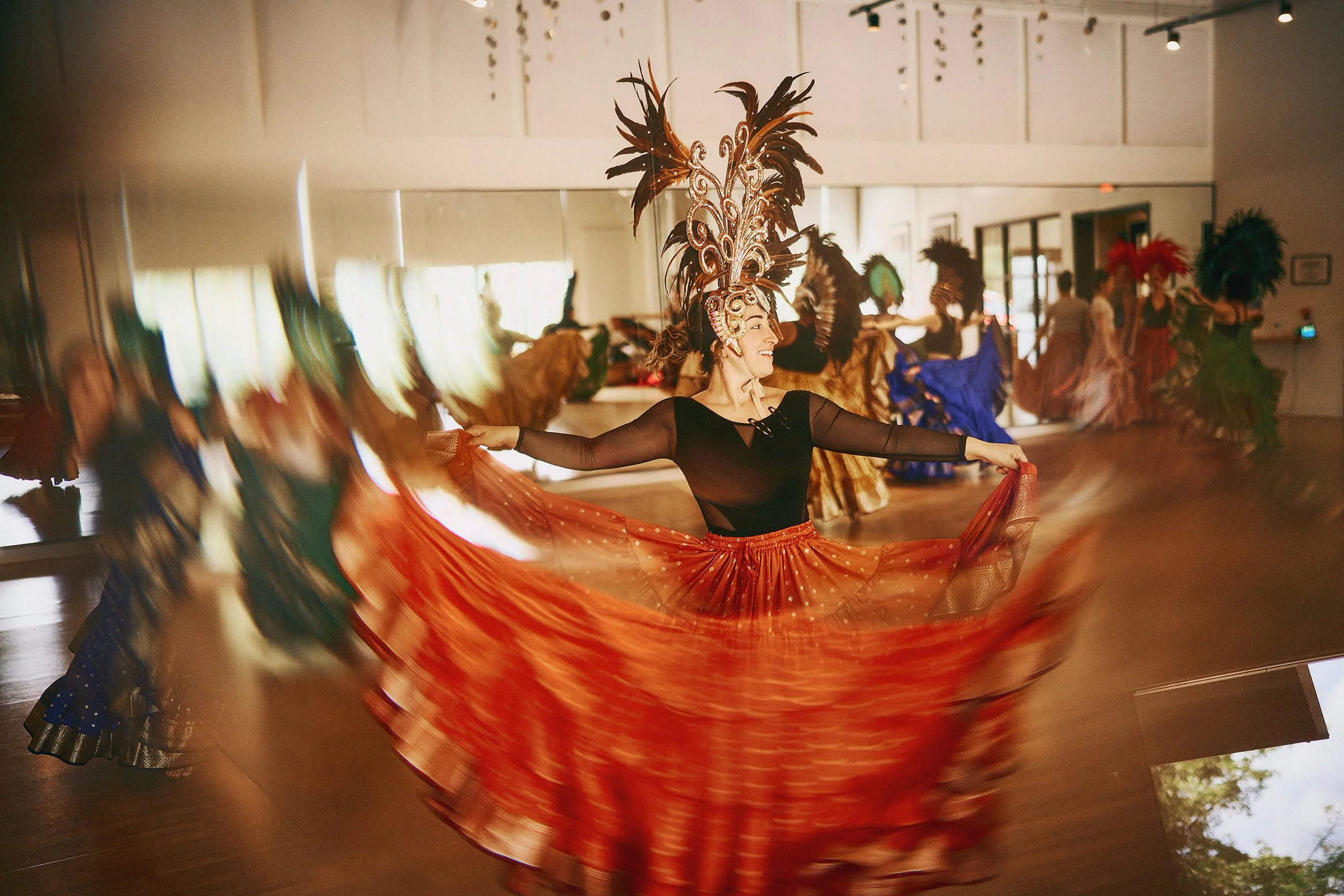 A woman performing a traditional dance in a room, wearing a large feathered headdress and holding a flowing orange skirt.