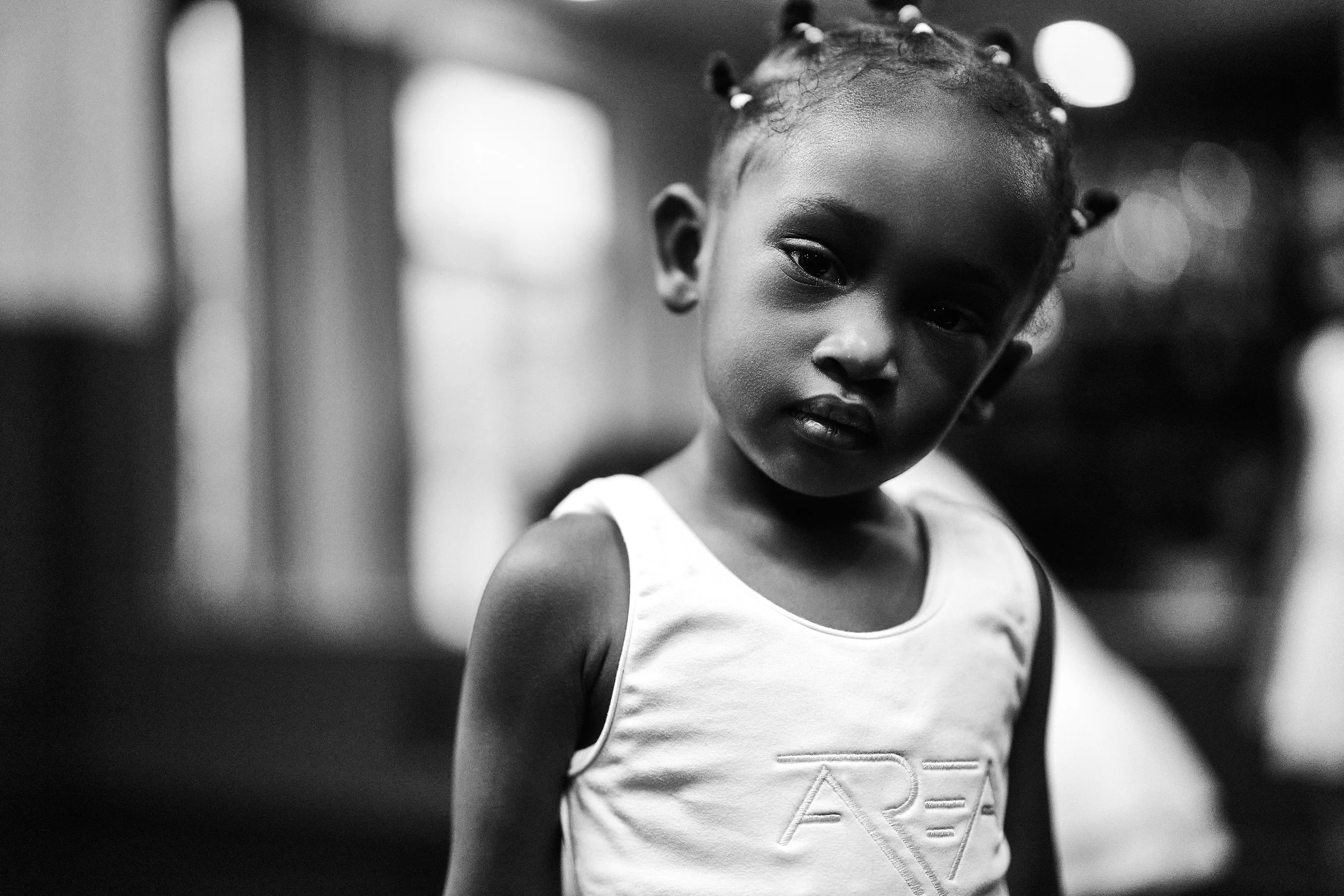 A young girl with braided hair and beads, wearing a sleeveless top, looking slightly downward in a black-and-white photo.