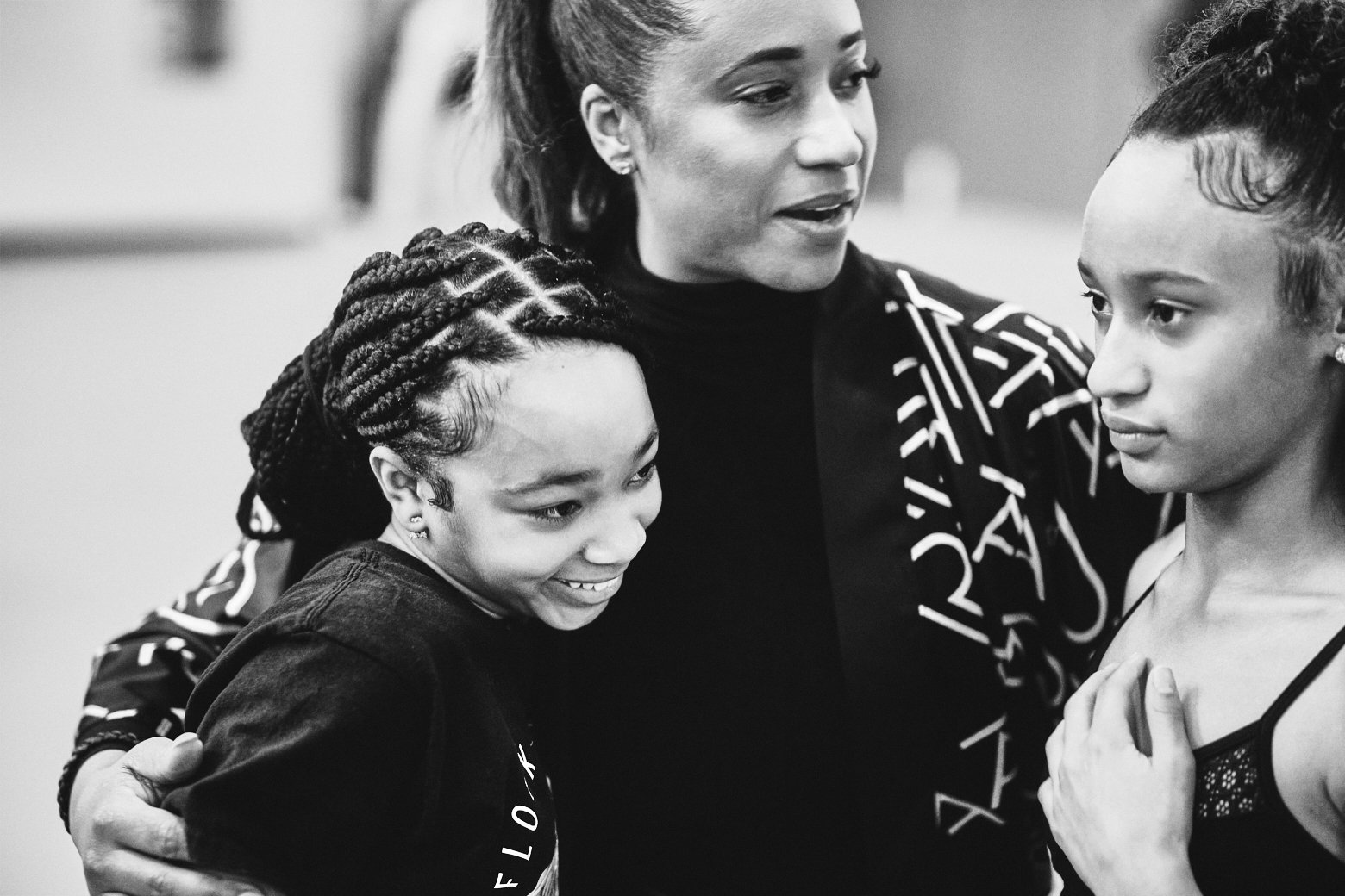 A woman with her hair pulled back speaks to two young girls. One girl has braids and smiles, while the other has curly hair and appears emotional. The woman is wearing a patterned jacket.