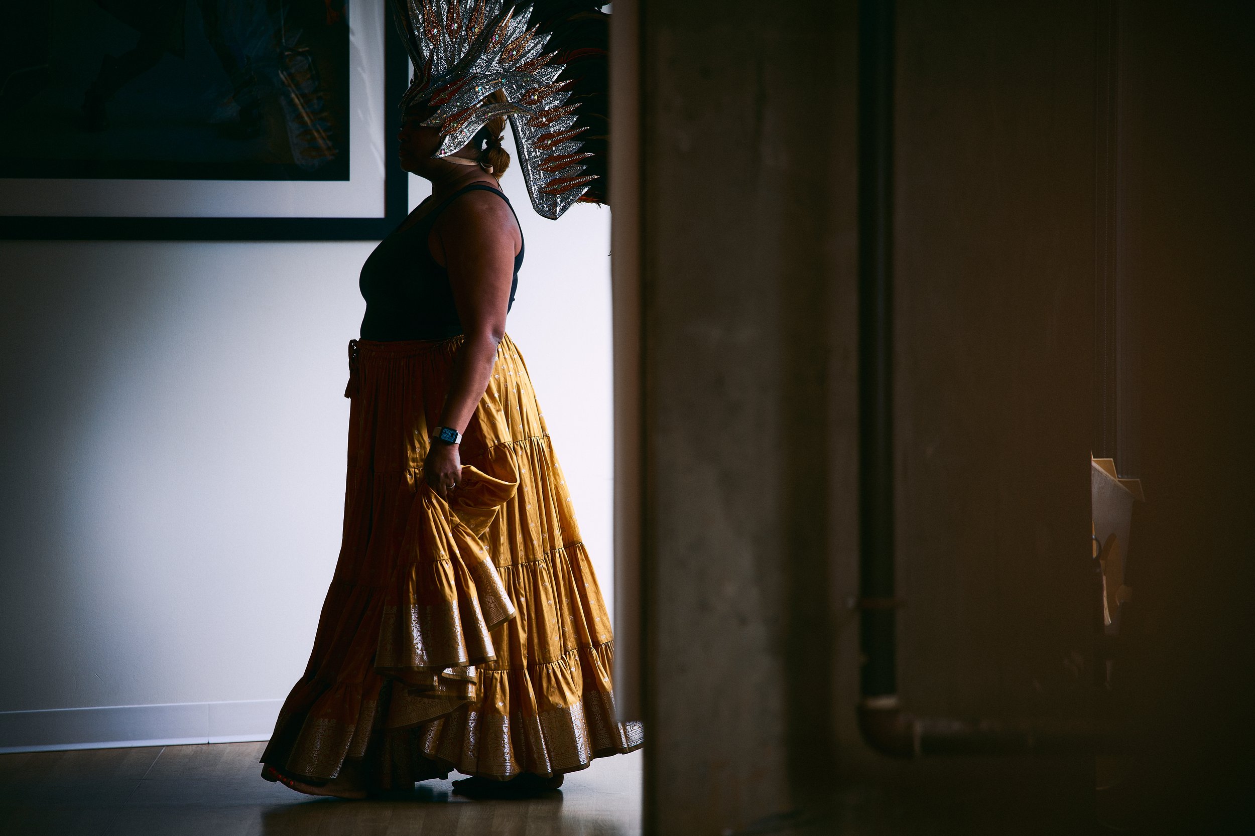 Person wearing elaborate feathered headdress and yellow skirt standing in a dimly lit room.