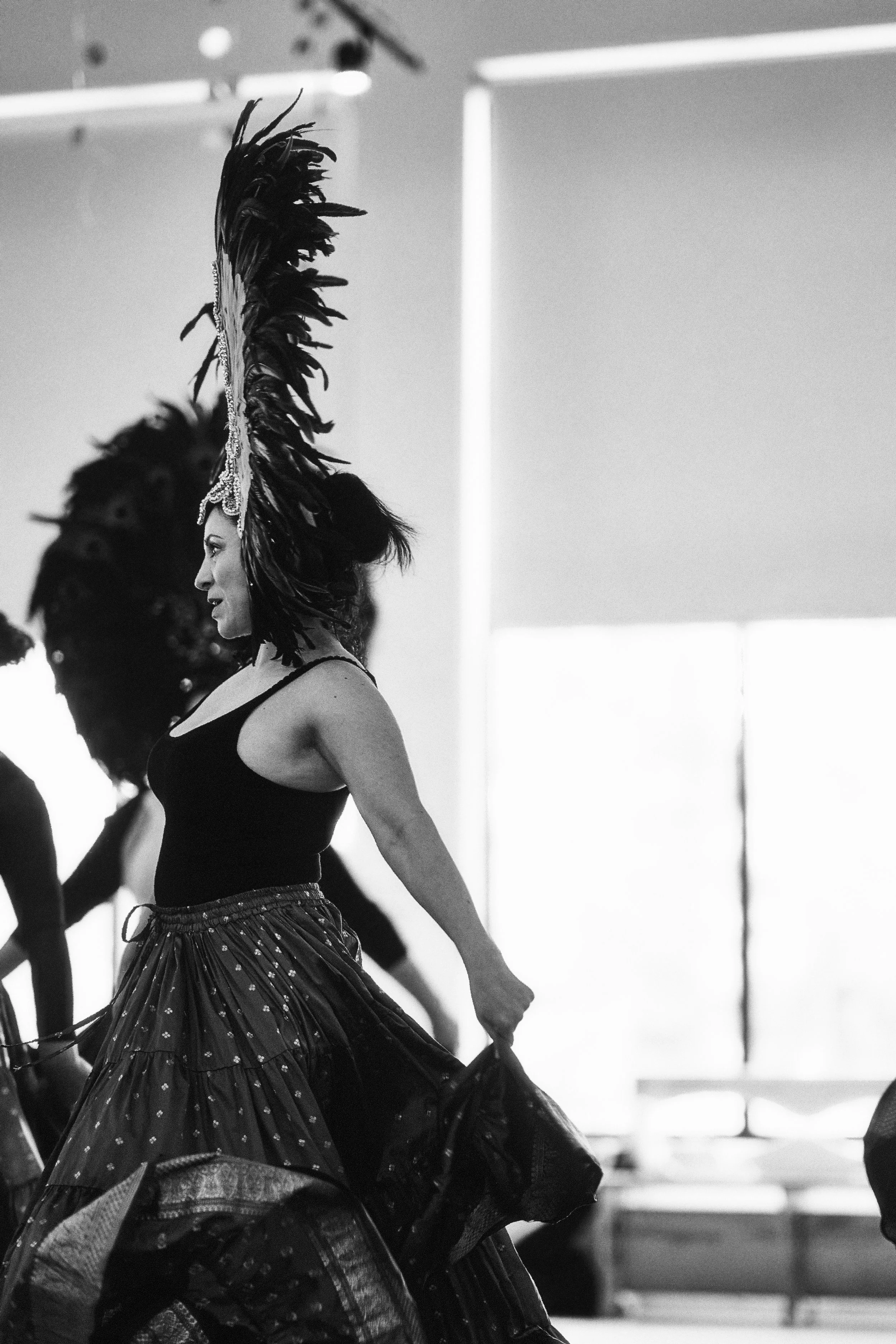 A woman dressed in a belly dance costume with a headpiece and feathered headdress, dancing in a studio or rehearsal space.