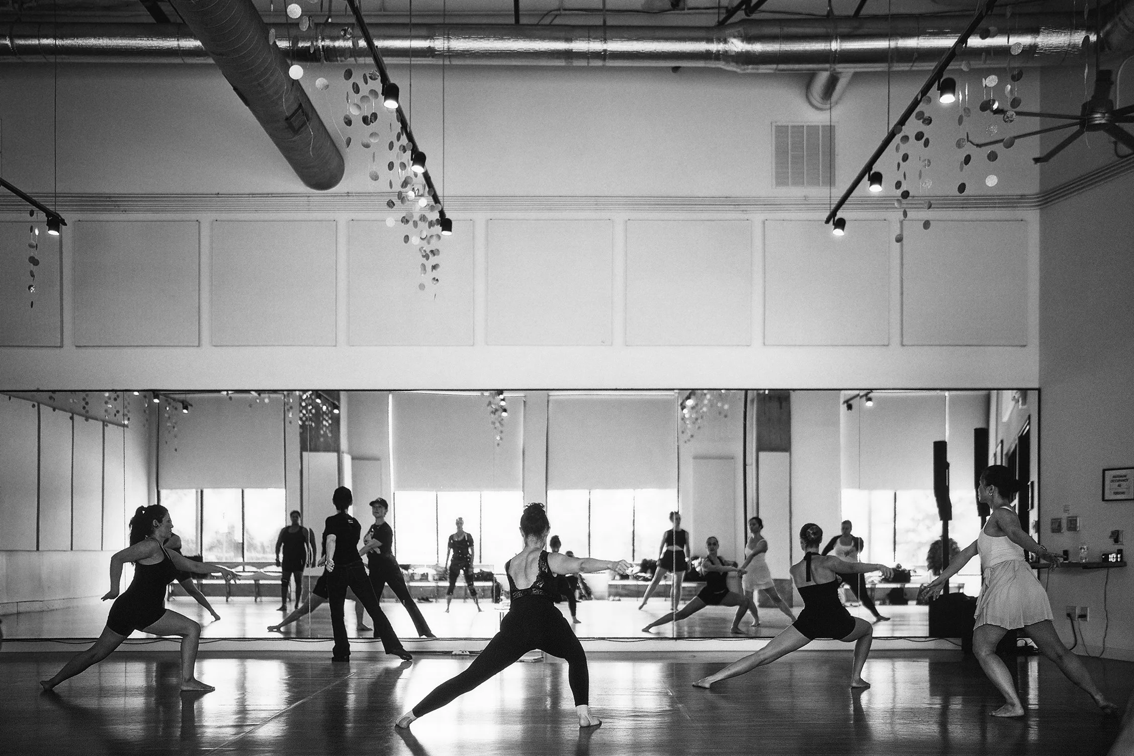 Ballet dancers practicing in a spacious dance studio with a mirrored wall and ventilation ducts on the ceiling.