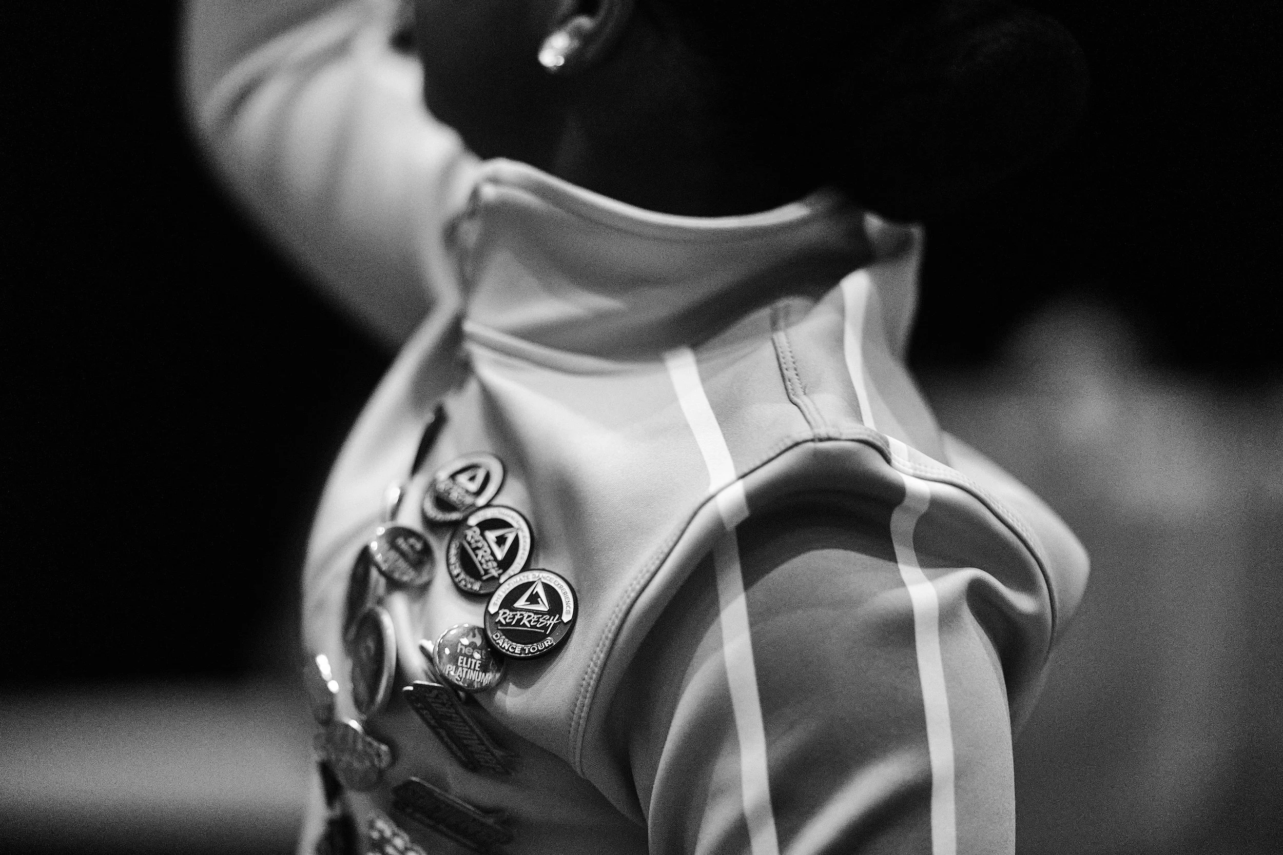 Close-up of a person wearing a jacket with multiple buttons, some of which have logos and text, including 'REPREZENT', 'DANCE TOUR', and 'ELITE PLATINUM'. The person's face is not visible.