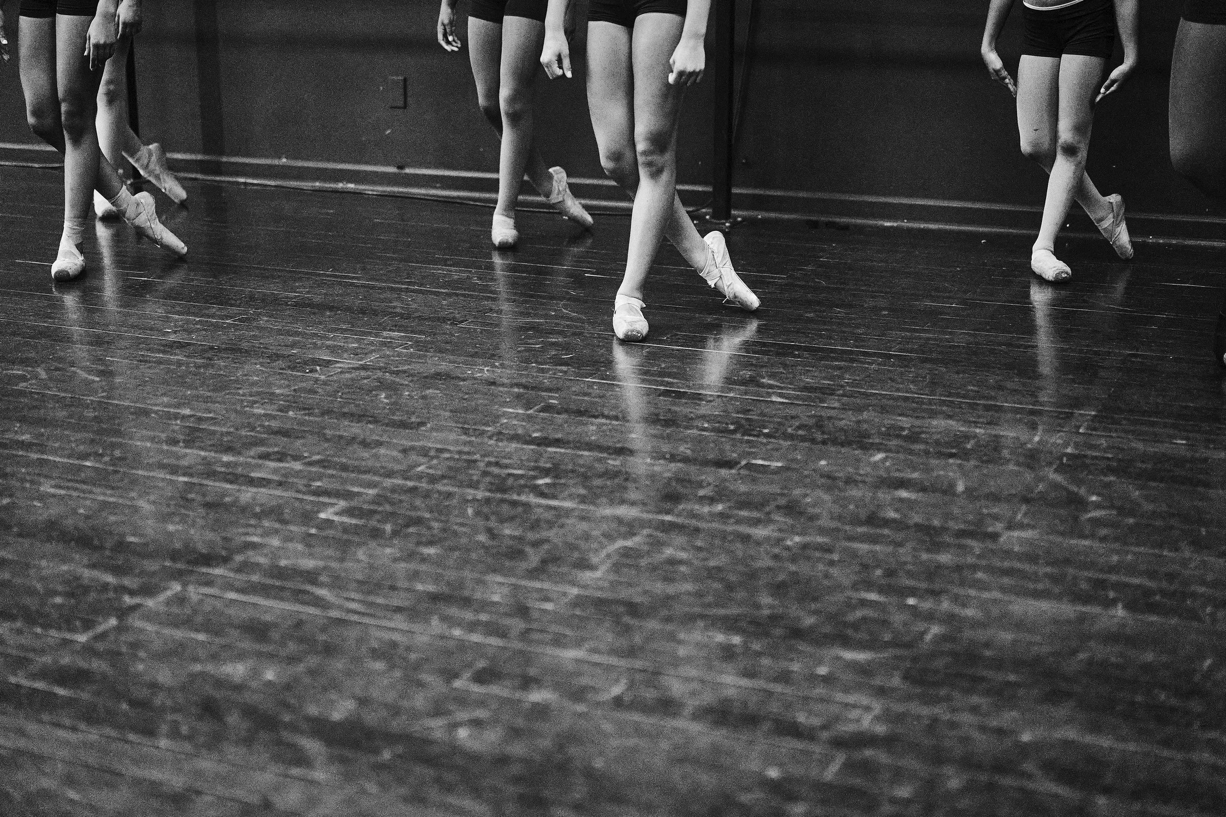 Black and white photo of ballet dancers' legs and feet, wearing pointe shoes, standing on a wooden dance floor against a dark wall.