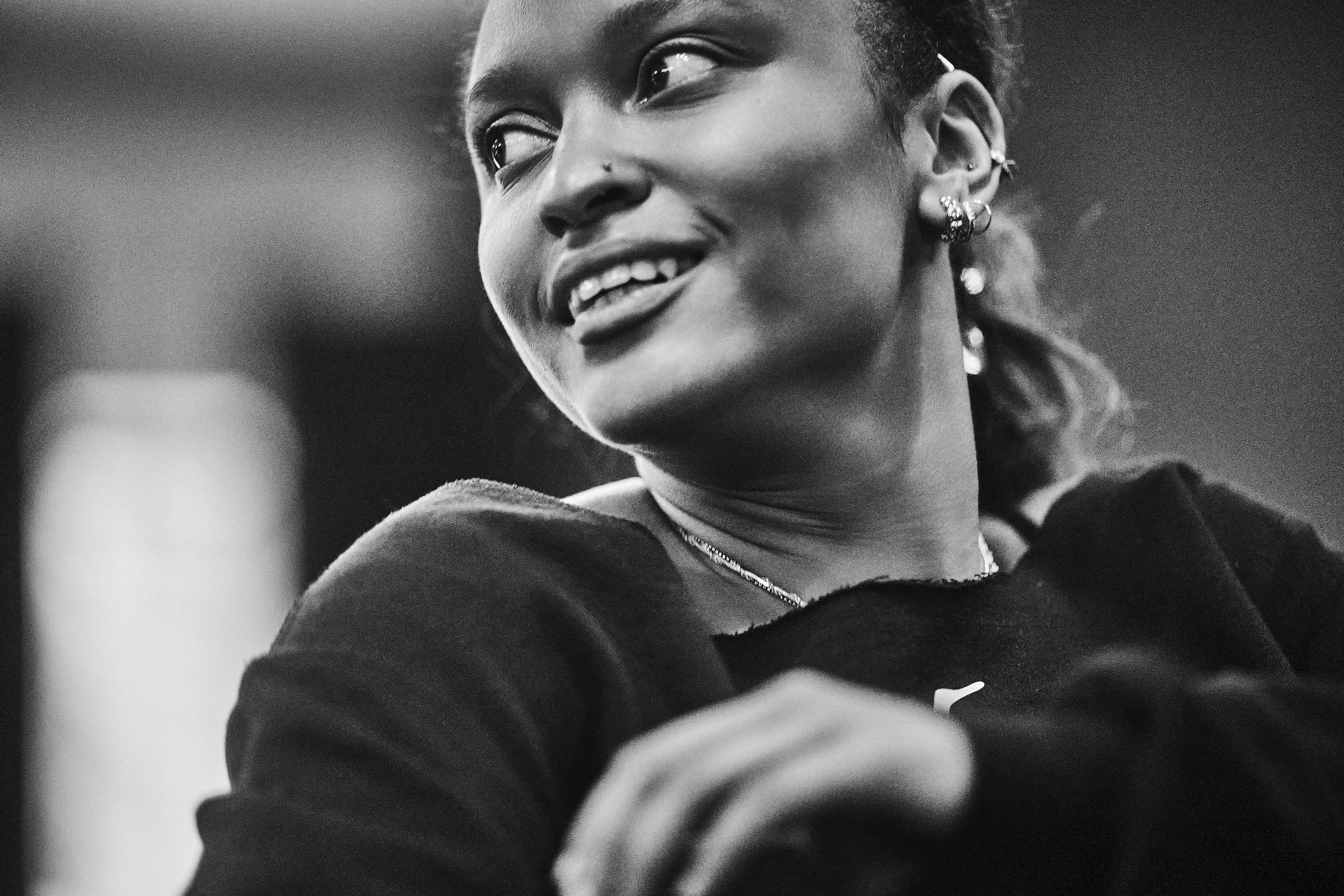 Close-up black and white photograph of a smiling woman with short hair, earrings, and layered necklaces, looking off to the side.