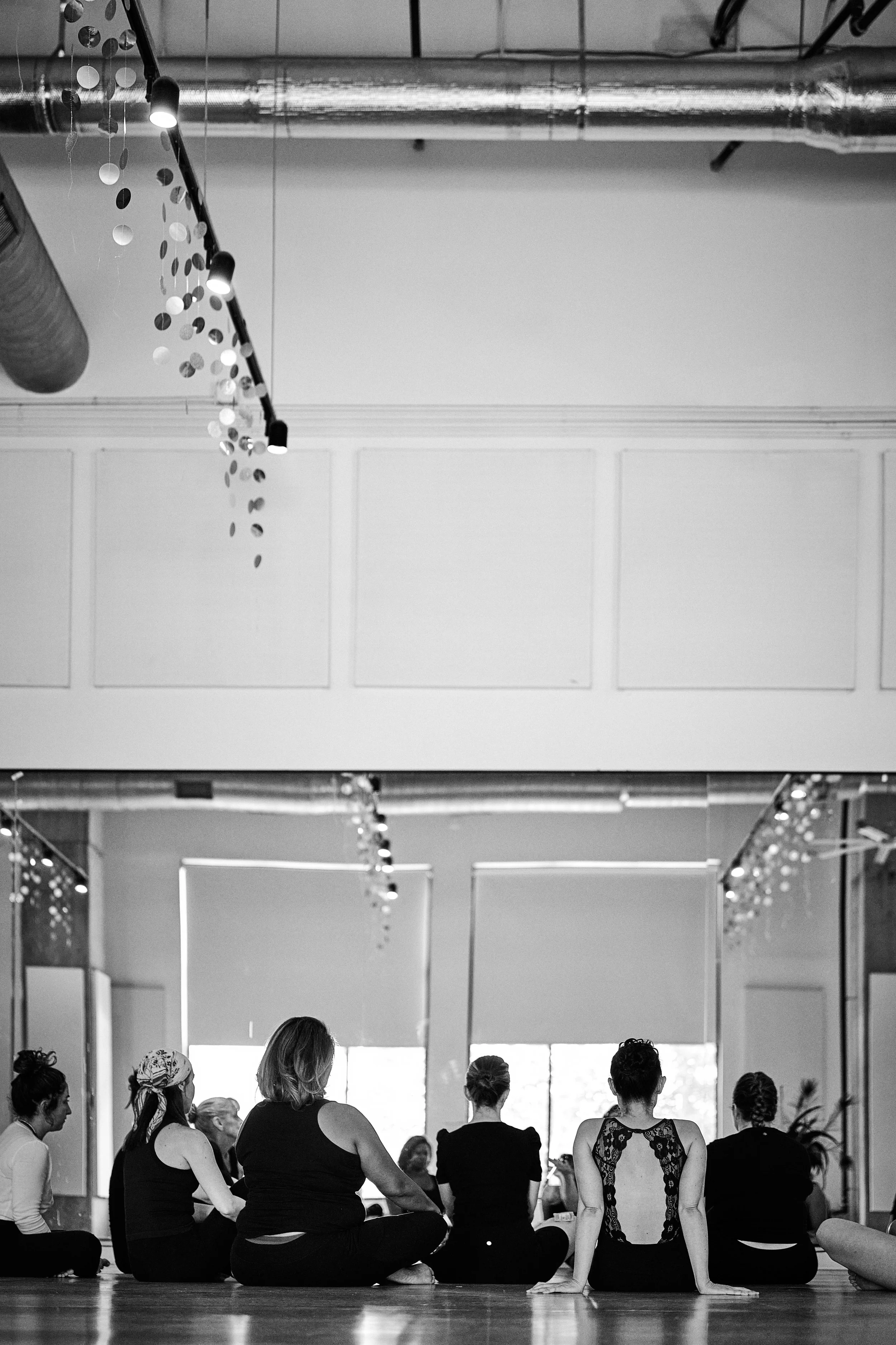 Group of women participating in a yoga class in a studio, sitting on the floor with their backs to the camera, facing a large window.