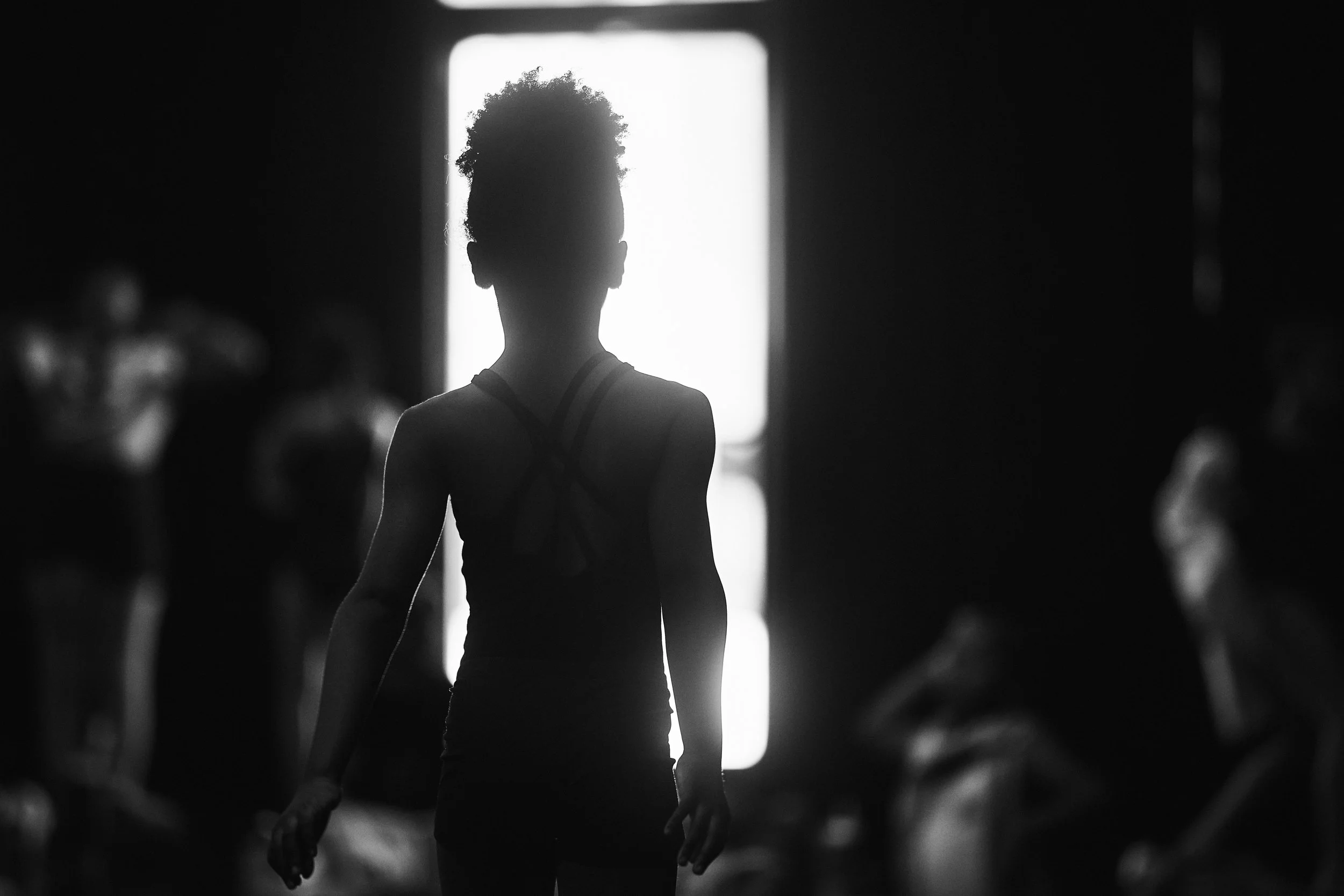 Silhouette of a woman with curly hair standing in front of a bright door, with blurred people seated in the background.