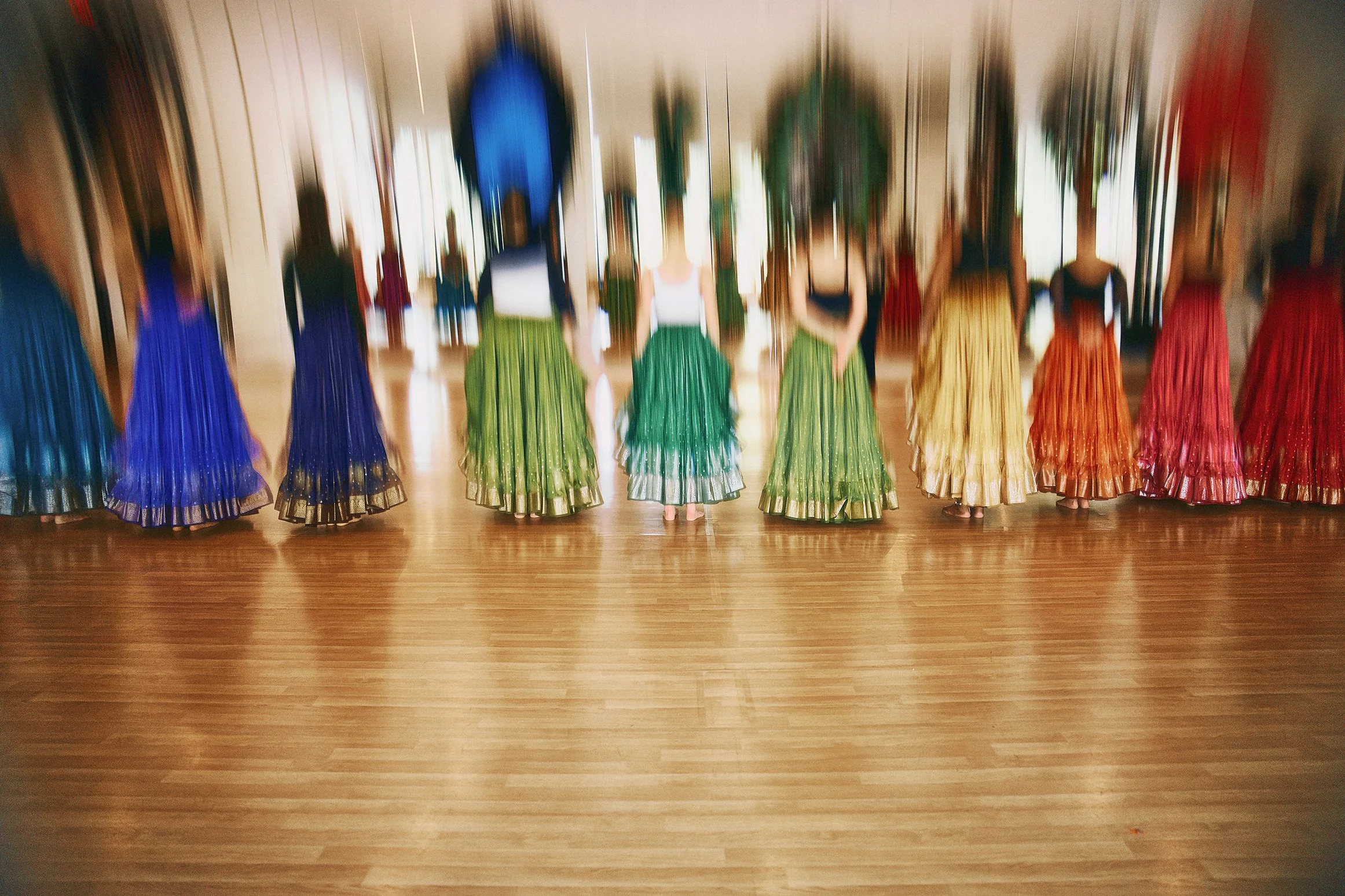 A group of women standing in a line wearing colorful, traditional skirts in shades of blue, green, yellow, orange, and red.
