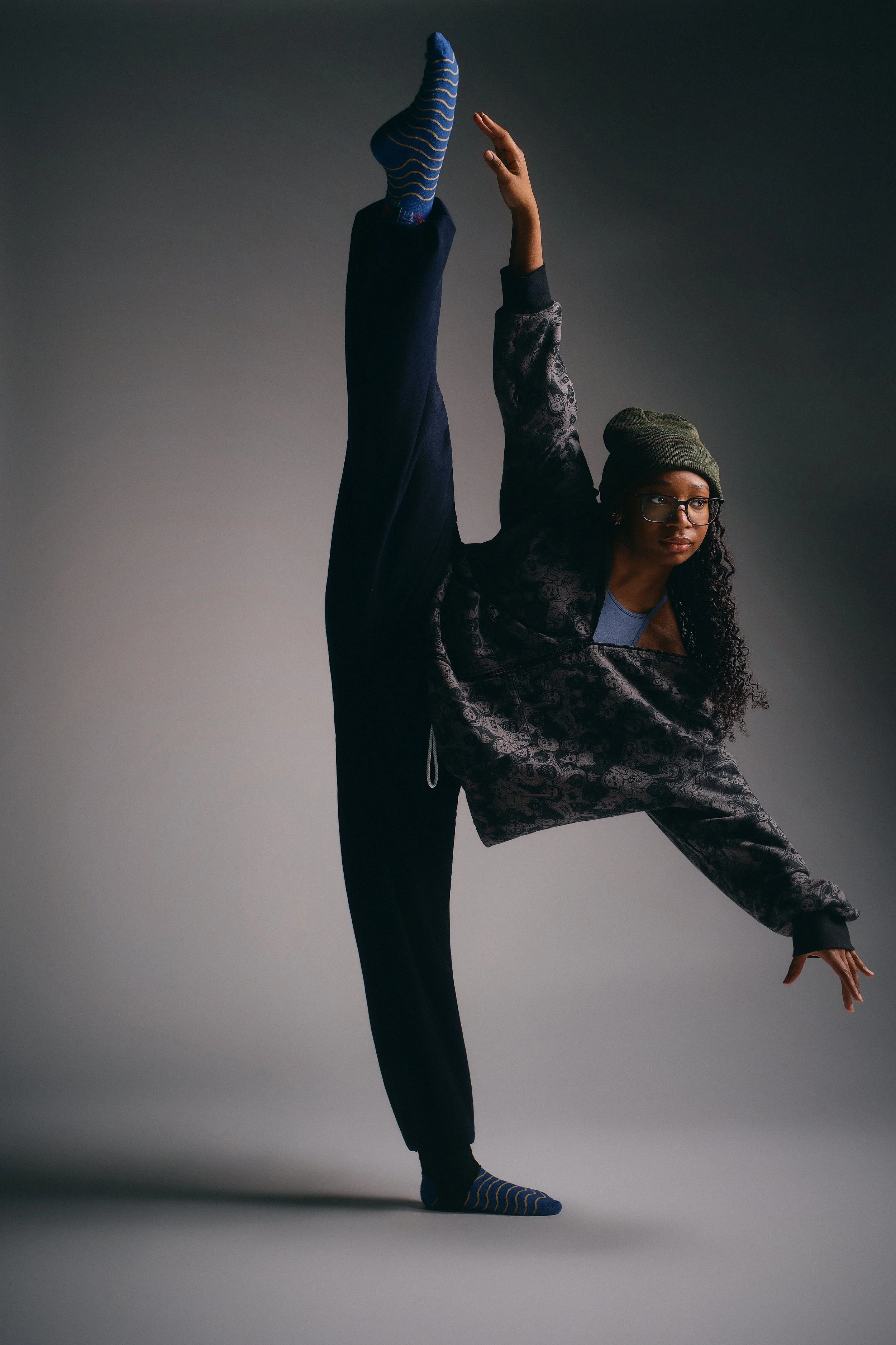 A woman with glasses and curly hair wearing a beanie, patterned jacket, and black pants performing a yoga or dance pose with one leg raised high and hand reaching towards the foot, balancing on her other leg.