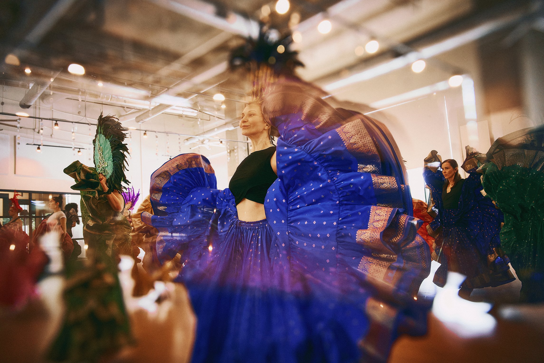 A woman in traditional Indian attire, holding hands with others, appears to be dancing during a cultural celebration or festival, with flowers in the foreground and decorated indoors.