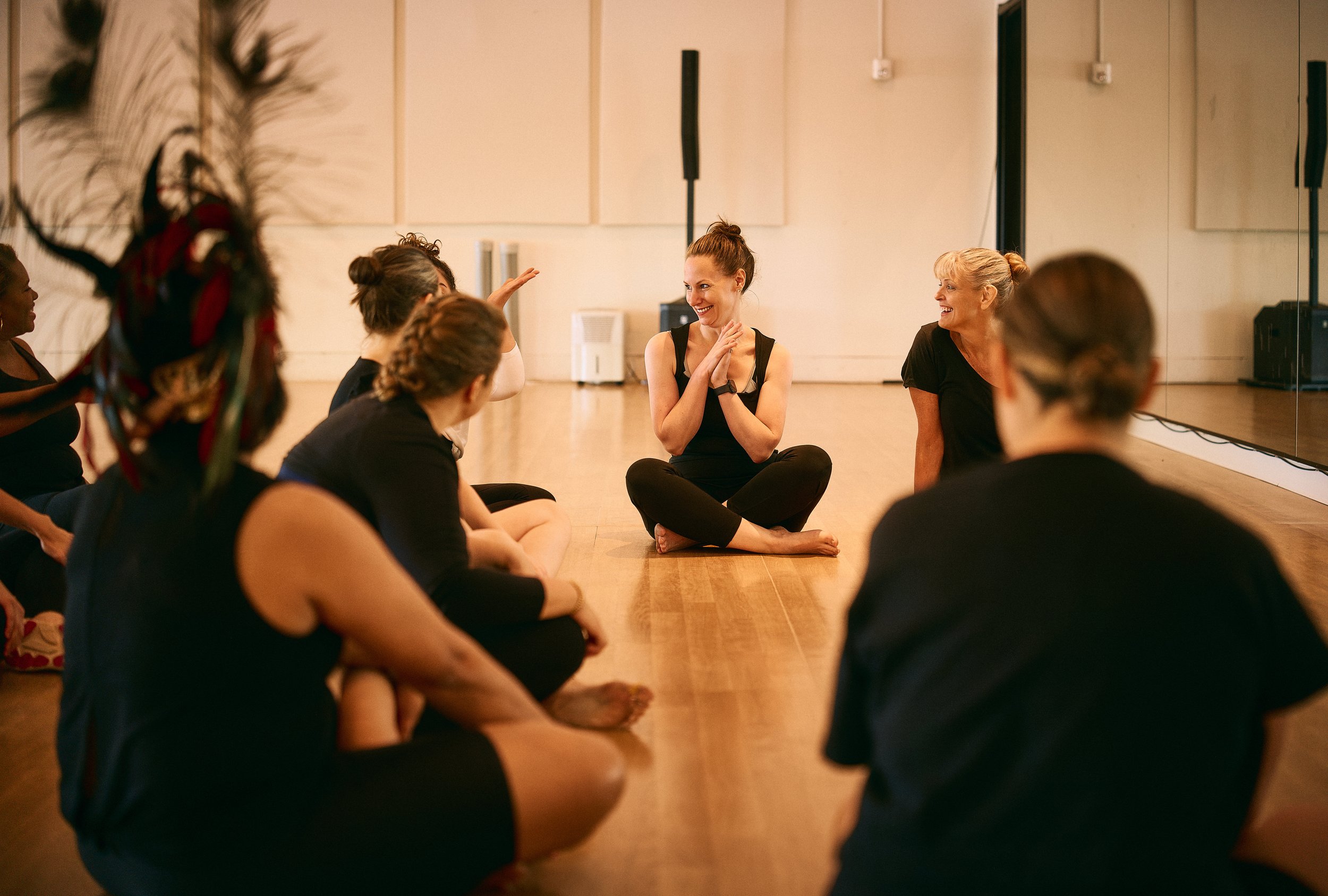 Group of women sitting on the floor of a dance studio, engaging in a discussion or workshop led by a woman in a black tank top, who is smiling and gesturing with her hands. The women appear attentive and involved in an activity.