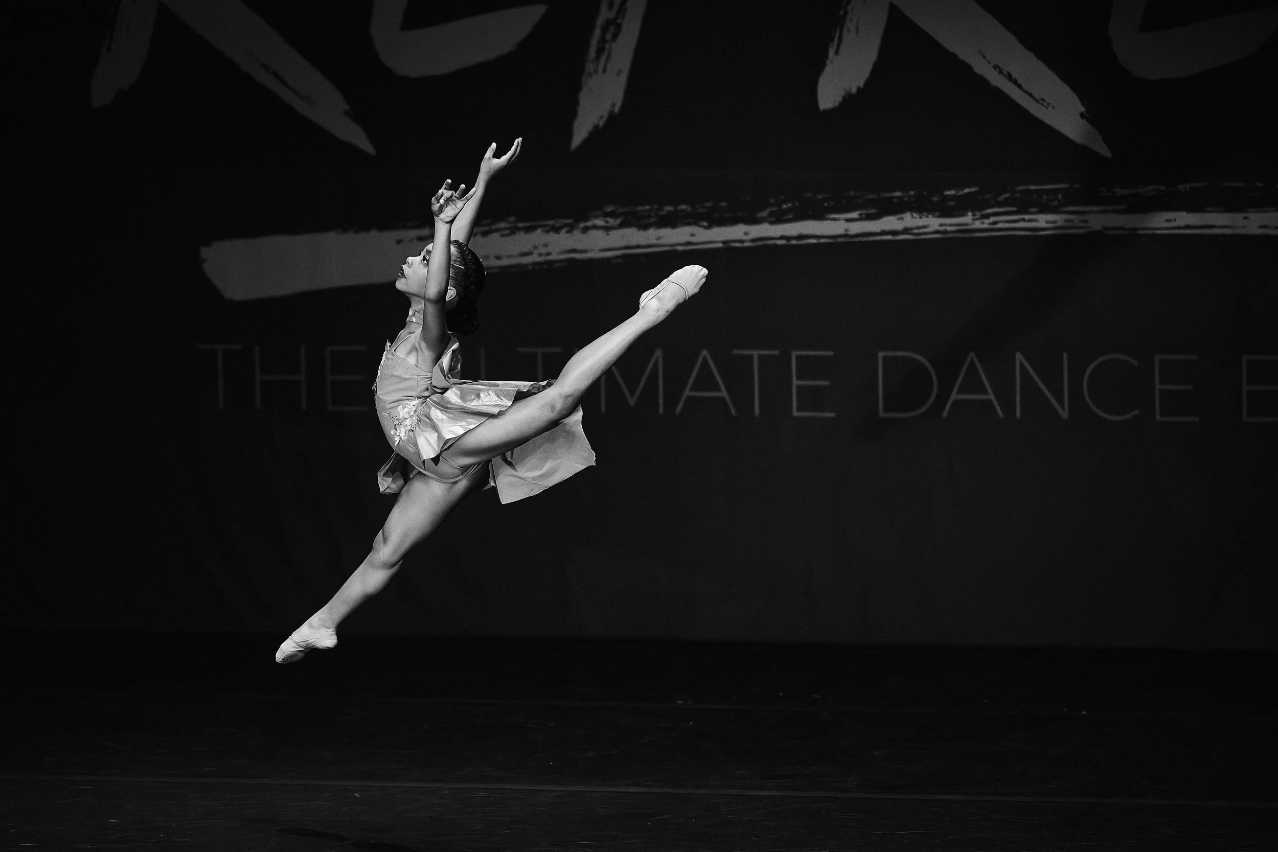 A ballet dancer performing an arabesque on stage with a dark background and a faint backdrop that reads 'The Ultimate Dance'.
