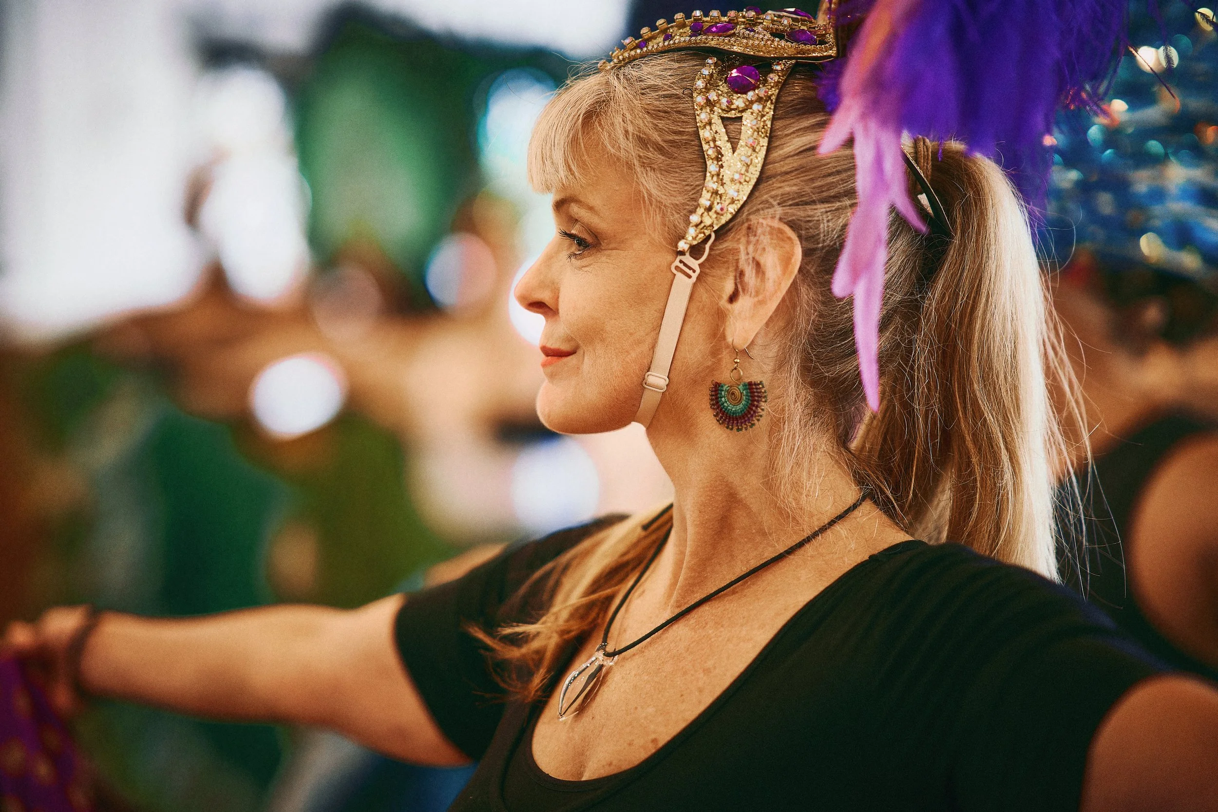 A woman with blonde hair tied back, wearing a shiny headband with purple gemstones, colorful earrings, and layered necklaces, appears to be dancing or performing at an event with colorful blurred lights in the background.