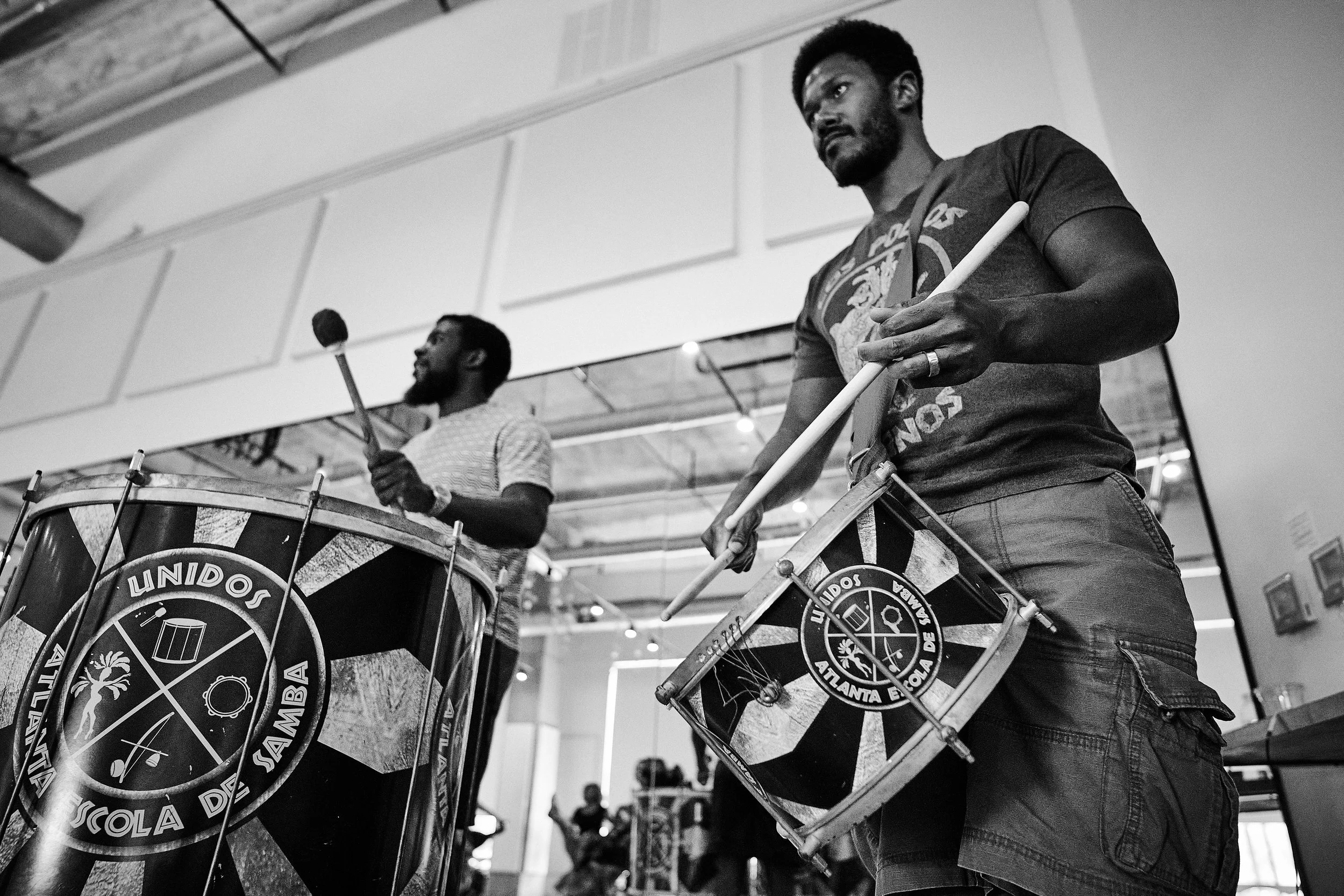 Two musicians playing drums at an indoor event. The drums are decorated with a circular logo featuring various icons and text. The man in the foreground is wearing a t-shirt and appears focused, holding drumsticks. The background is blurred with visible ceiling and some people.