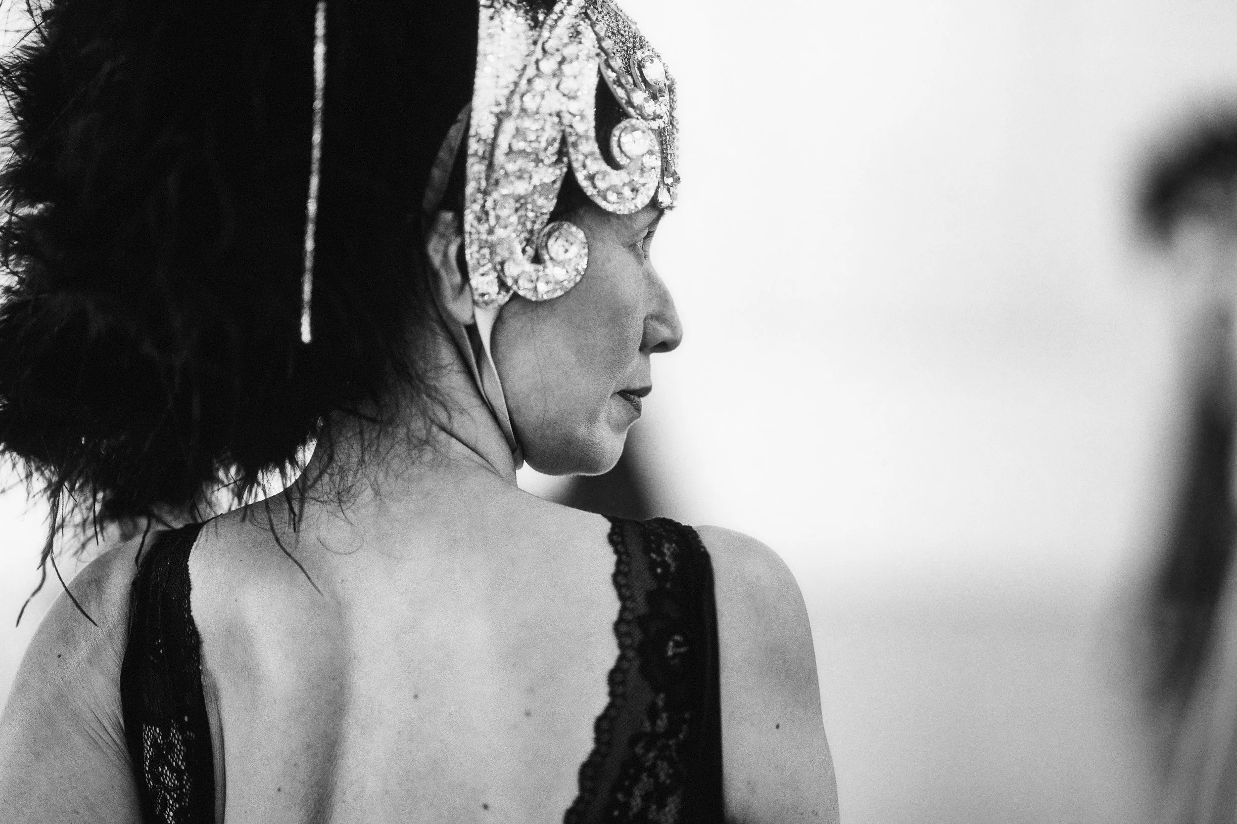 A black and white photo of a woman with dark hair styled in an updo, wearing a bejeweled headpiece and a lace dress with thin straps.