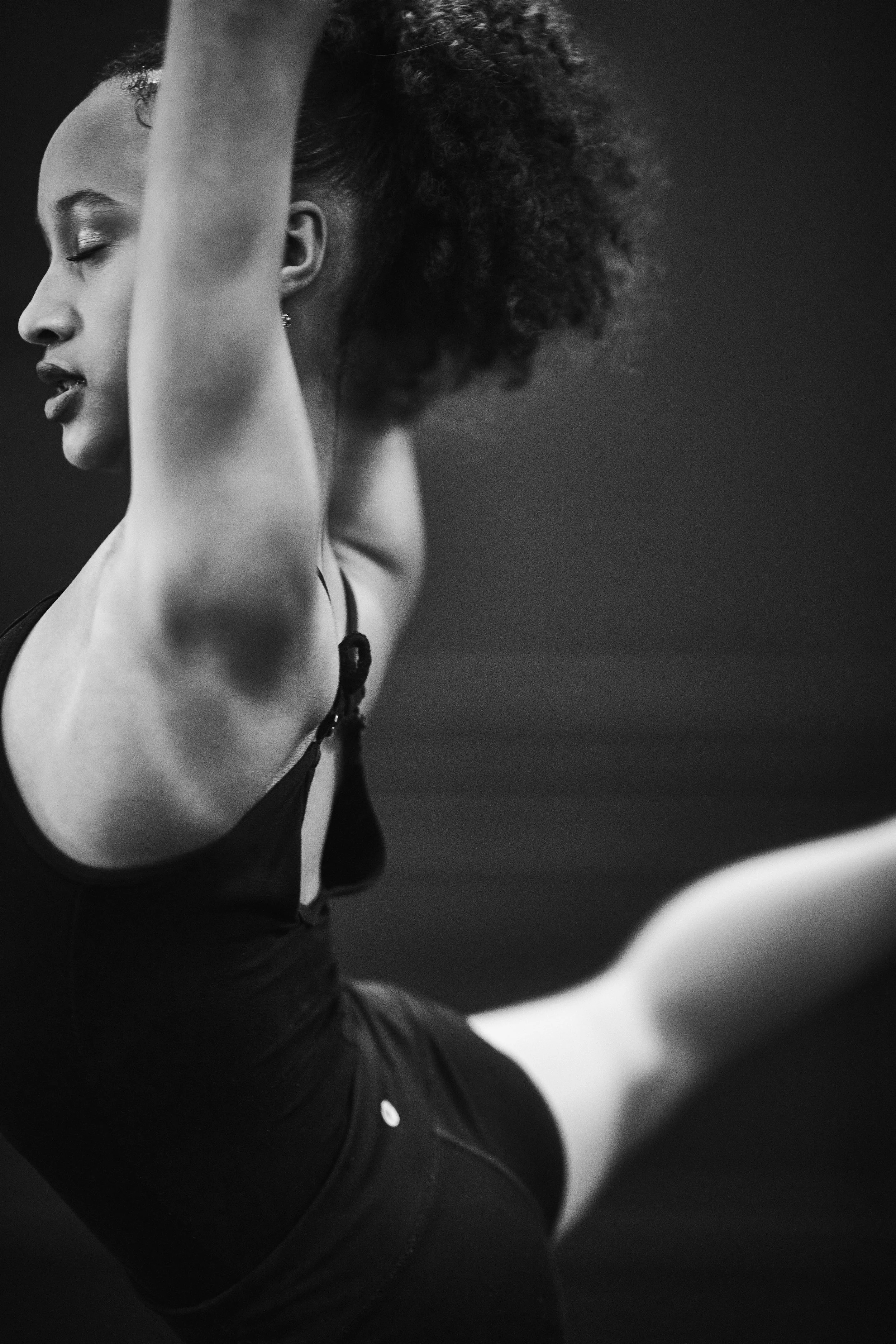 A Black woman in a black athletic outfit stretching or exercising indoors, with her arms raised above her head and her eyes closed.