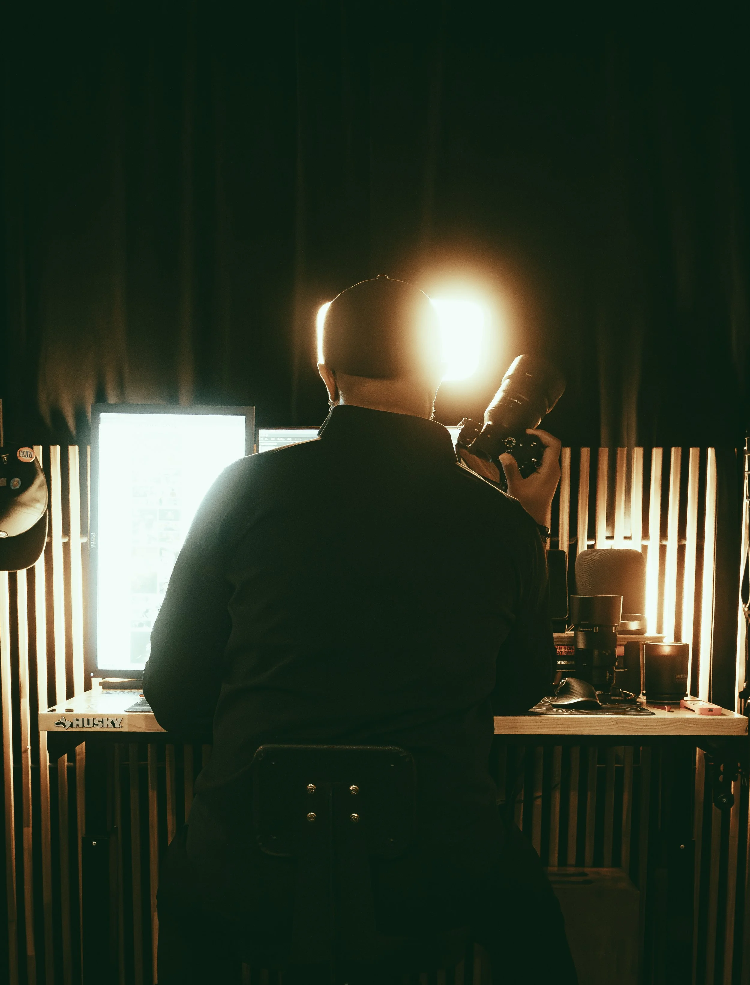 Person seated at a workstation with a computer screen and camera equipment.