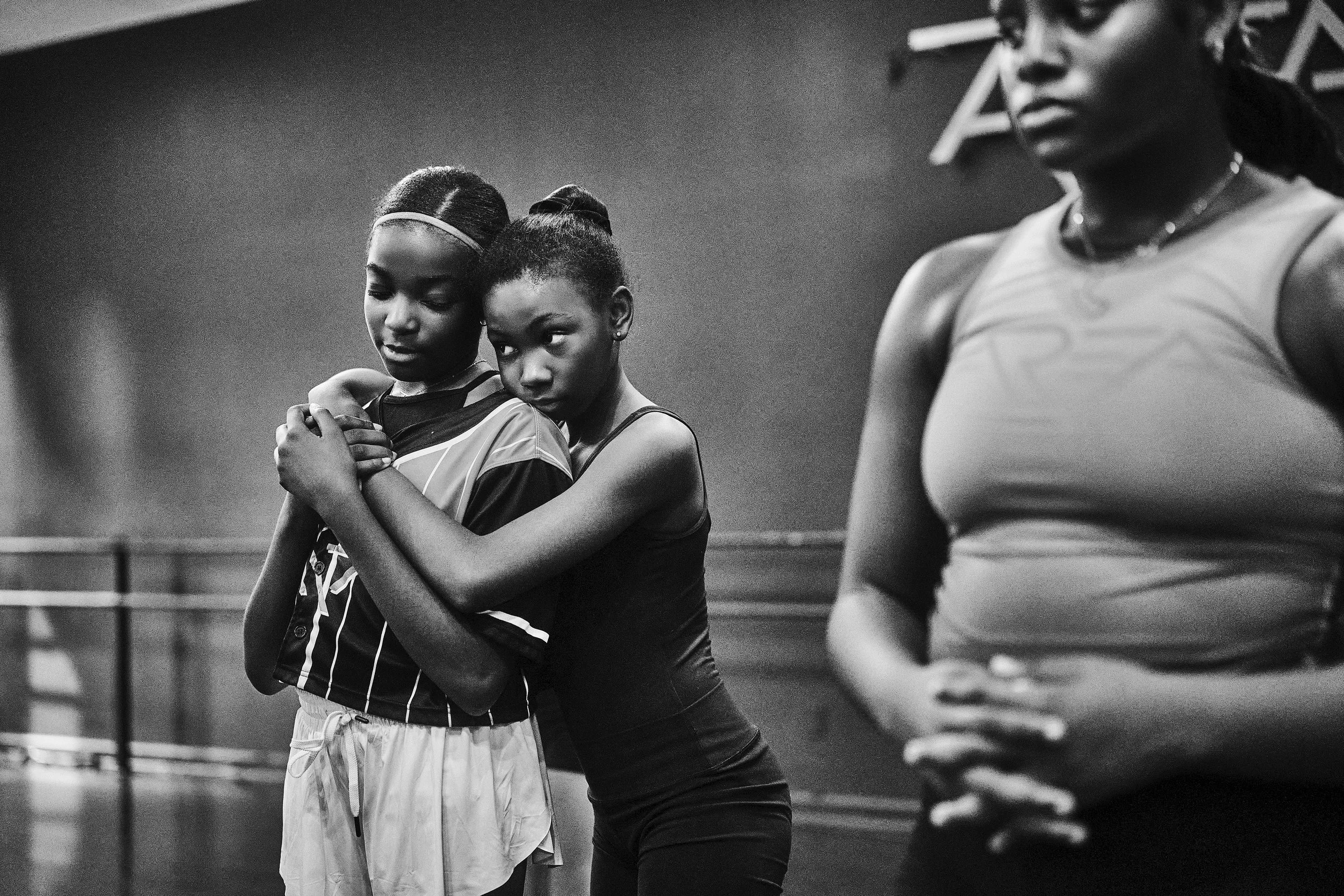 Two female athletes hugging during a sports event, with a third woman partially visible in the foreground. The photo is in black and white.