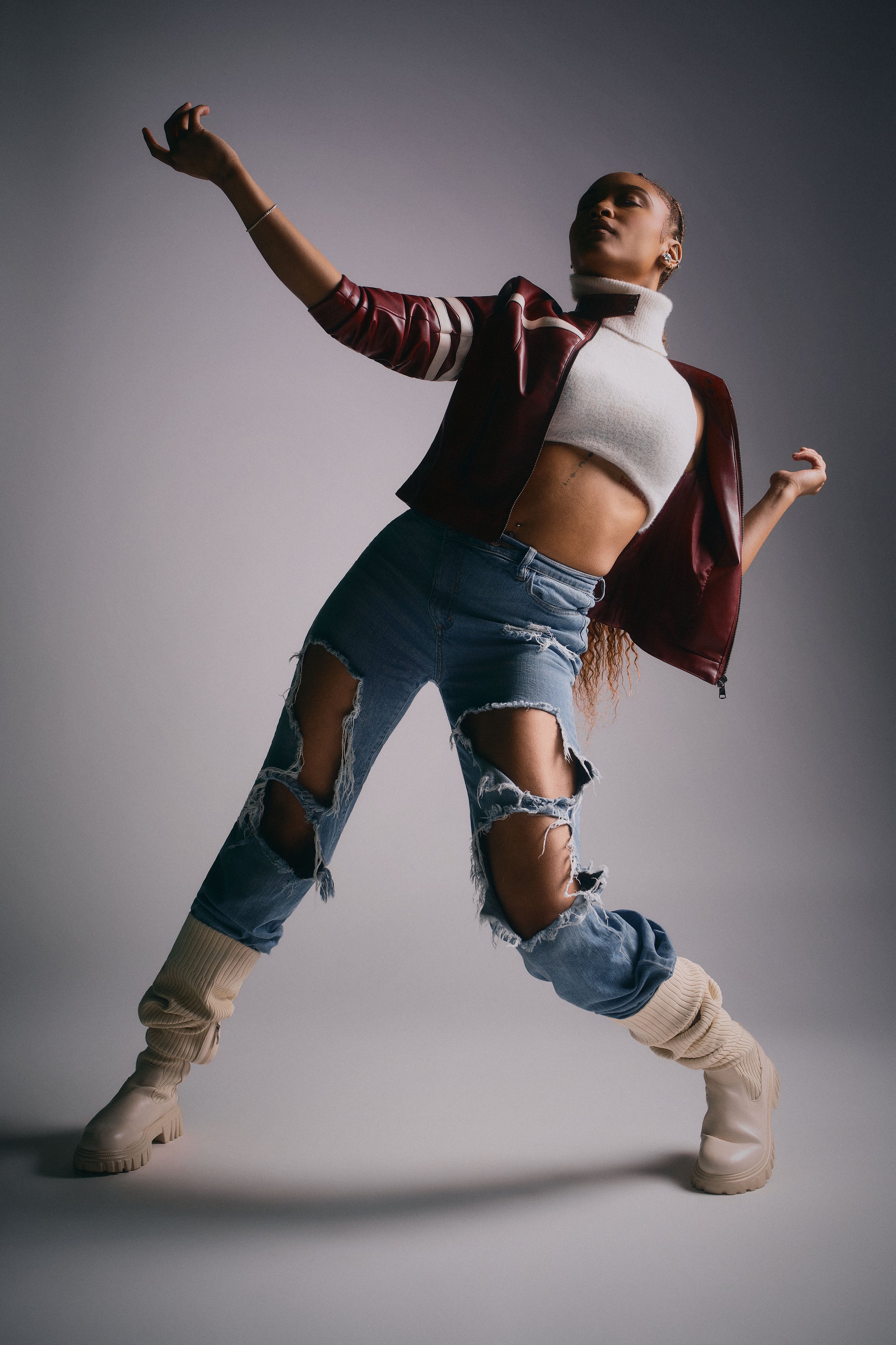 A woman in a pose with her arms raised and feet apart, wearing ripped jeans, a cropped top, a burgundy jacket, beige boots, and styled hair, standing against a plain background.